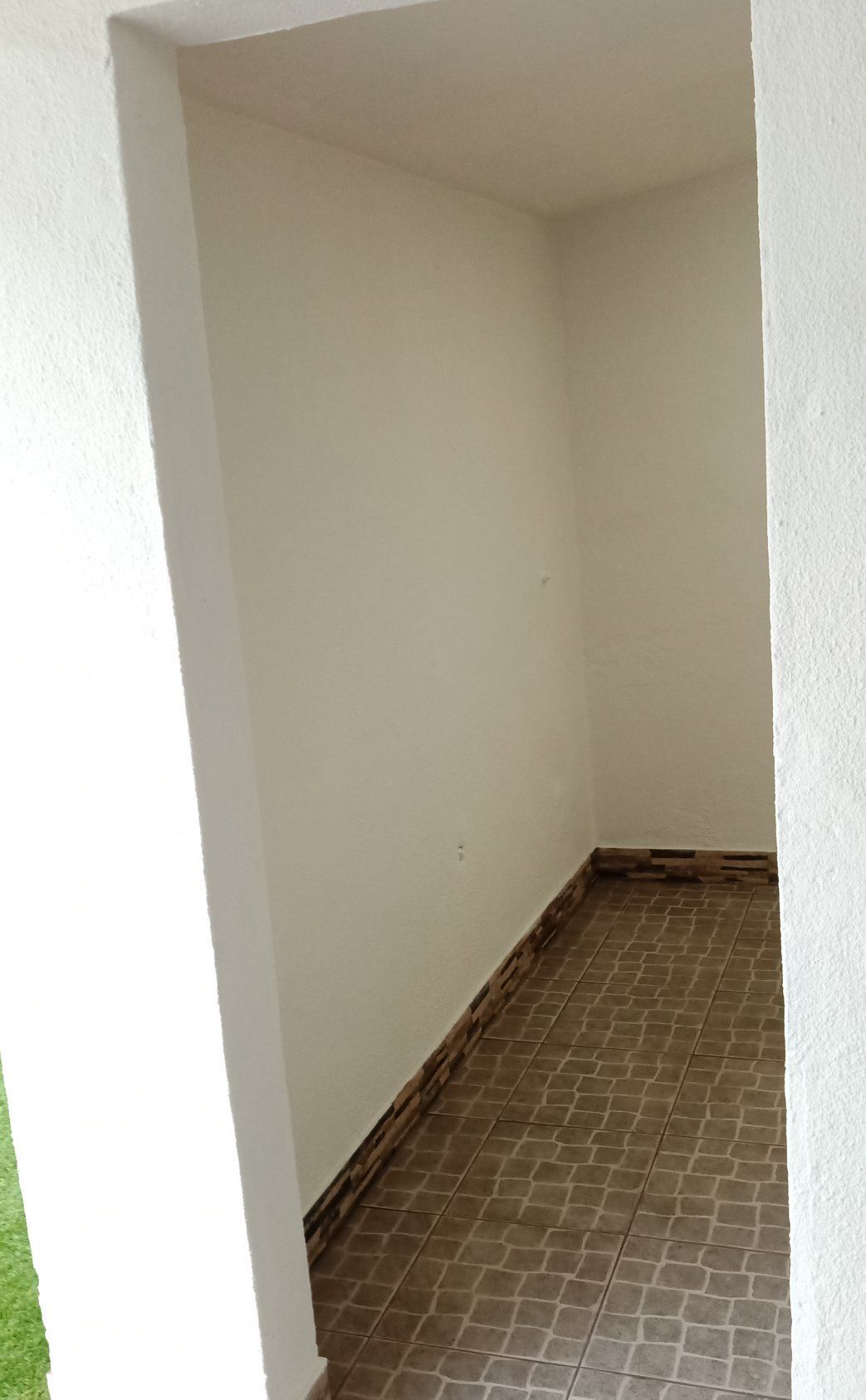 Quarto pequeno e vazio com paredes branco-sujas, piso de azulejo e uma porta à esquerda.