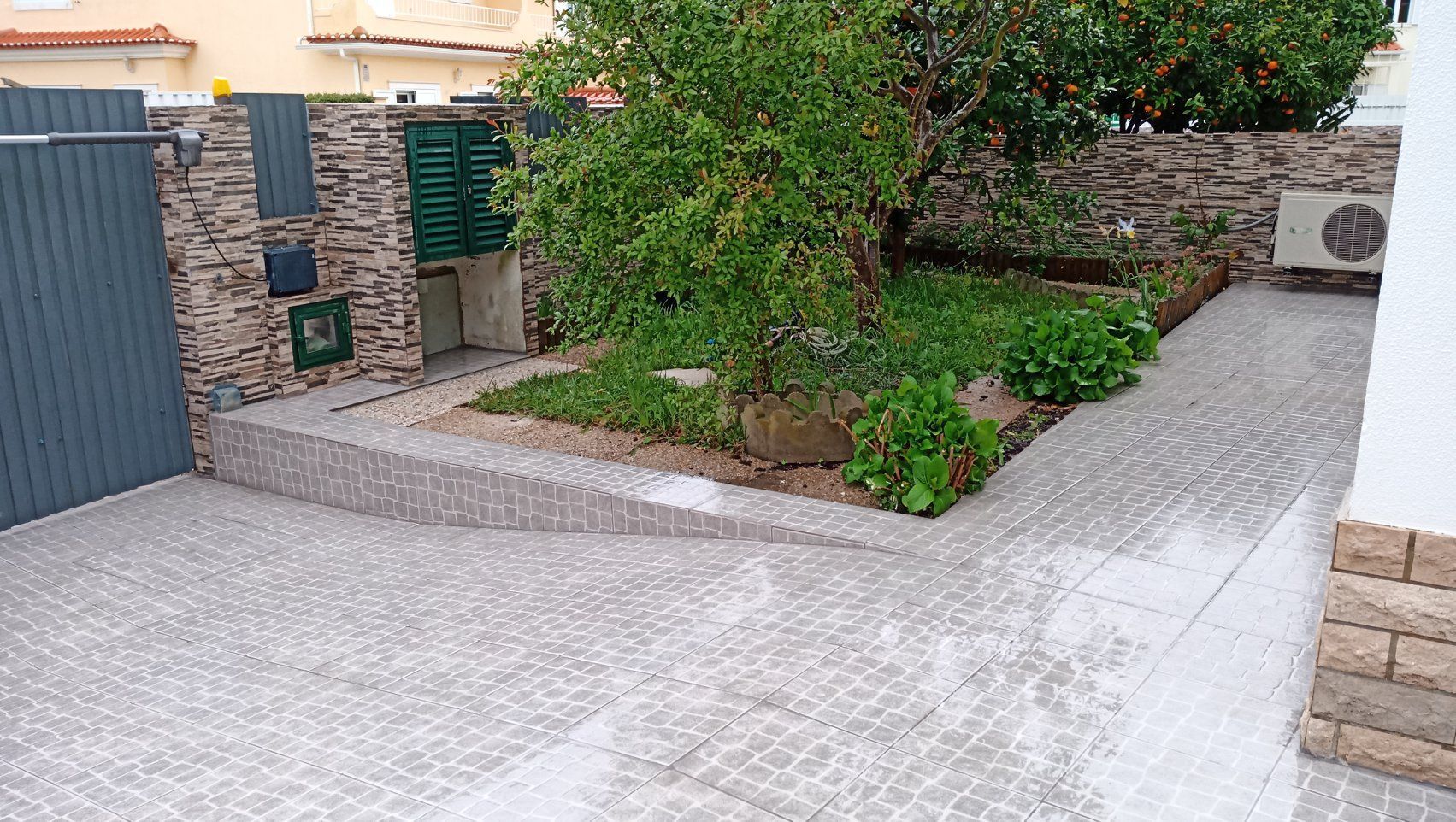 Entrada de automóveis pavimentada com paralelepípedos que leva a um pequeno jardim com uma árvore e um muro de pedra.