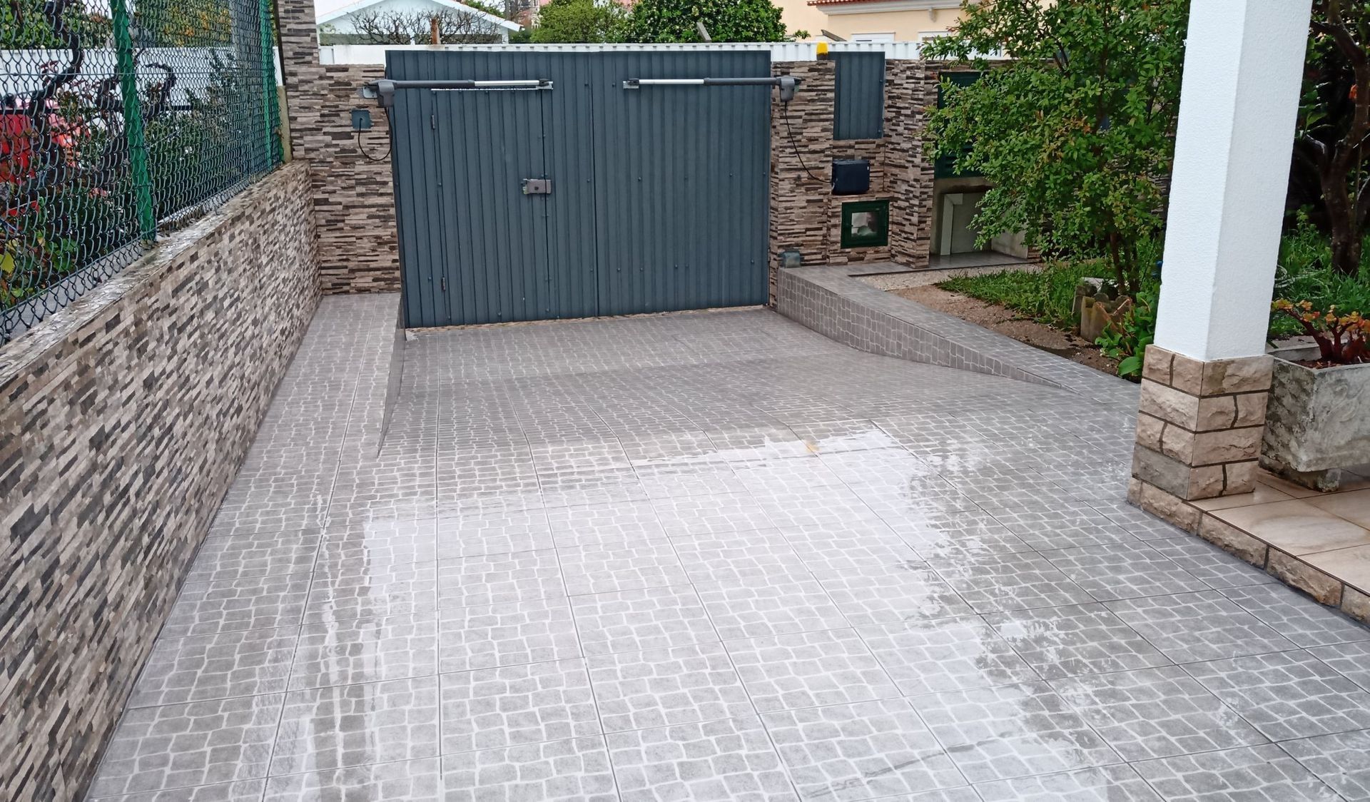 Entrada de automóveis em paralelepípedos cinzentos que leva a um portão cinzento fechado. Muros de pedra e um pilar branco são visíveis.