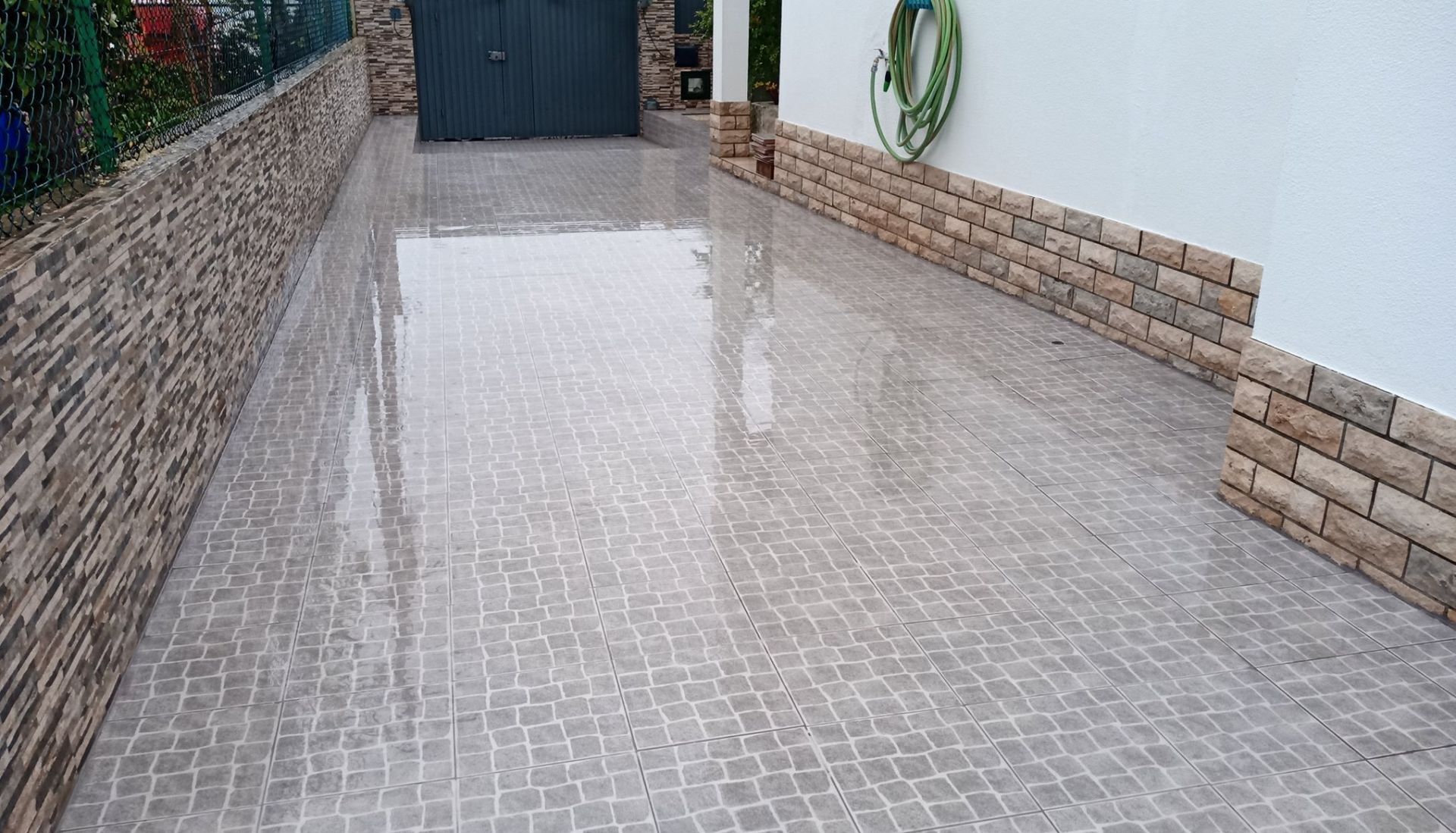 Calçada de paralelepípedos molhados que leva a uma porta de garagem escura, ladeada por paredes de tijolo e pedra.