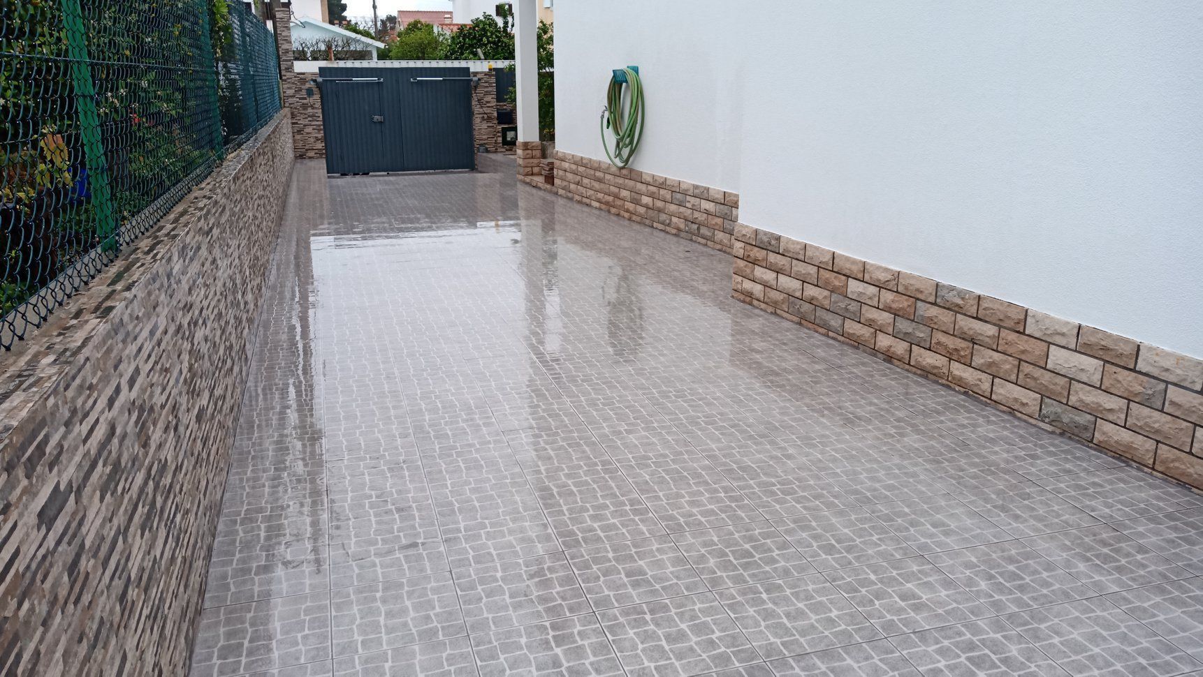 Entrada de automóveis molhada e revestida com azulejos, que leva a um portão preto fechado, delimitado por tijolos e um muro branco, com uma cerca verde à esquerda.