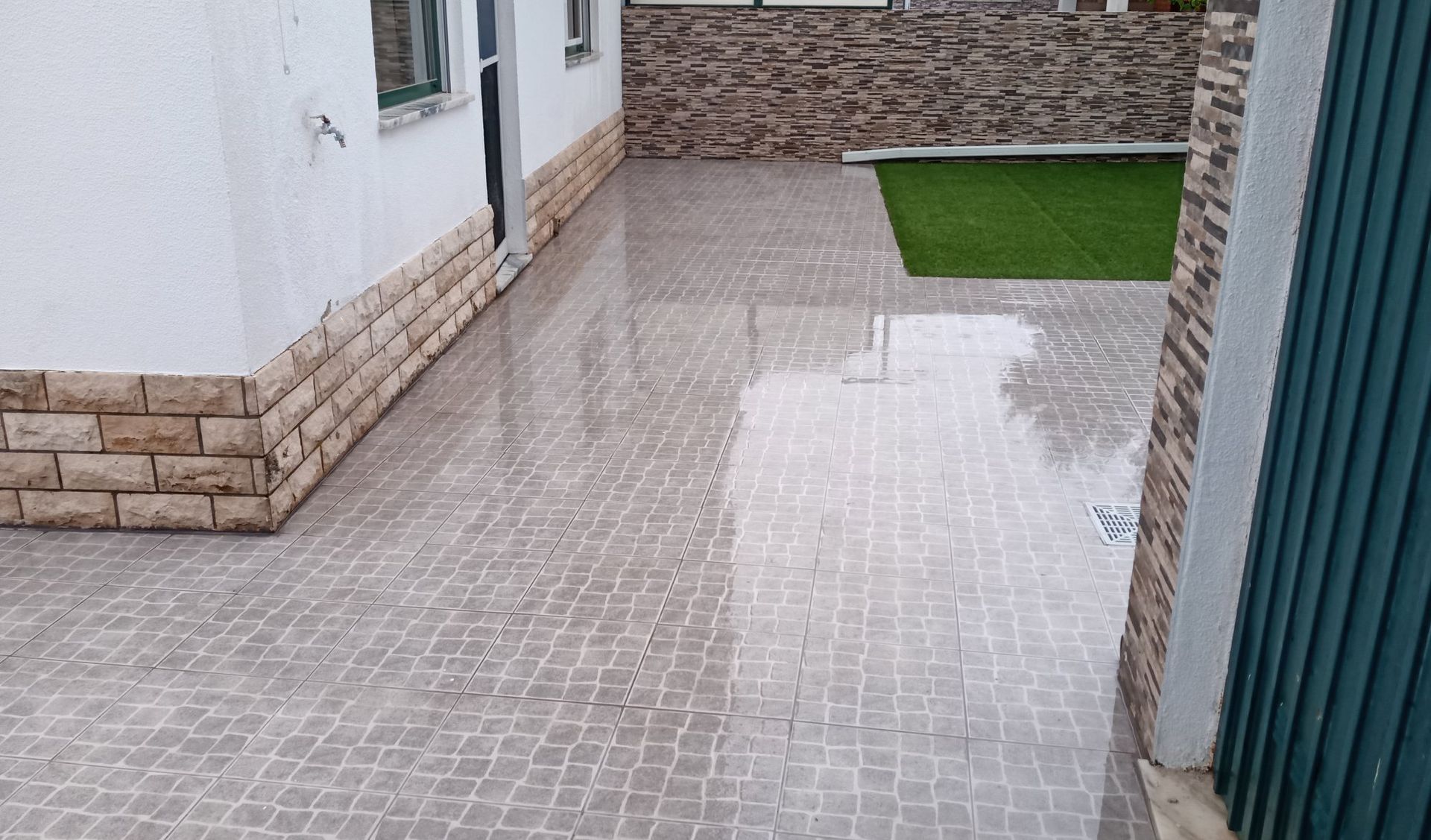 Pátio molhado com azulejos cinzentos ao lado de uma casa branca com detalhes em tijolo e uma área de grama artificial verde.