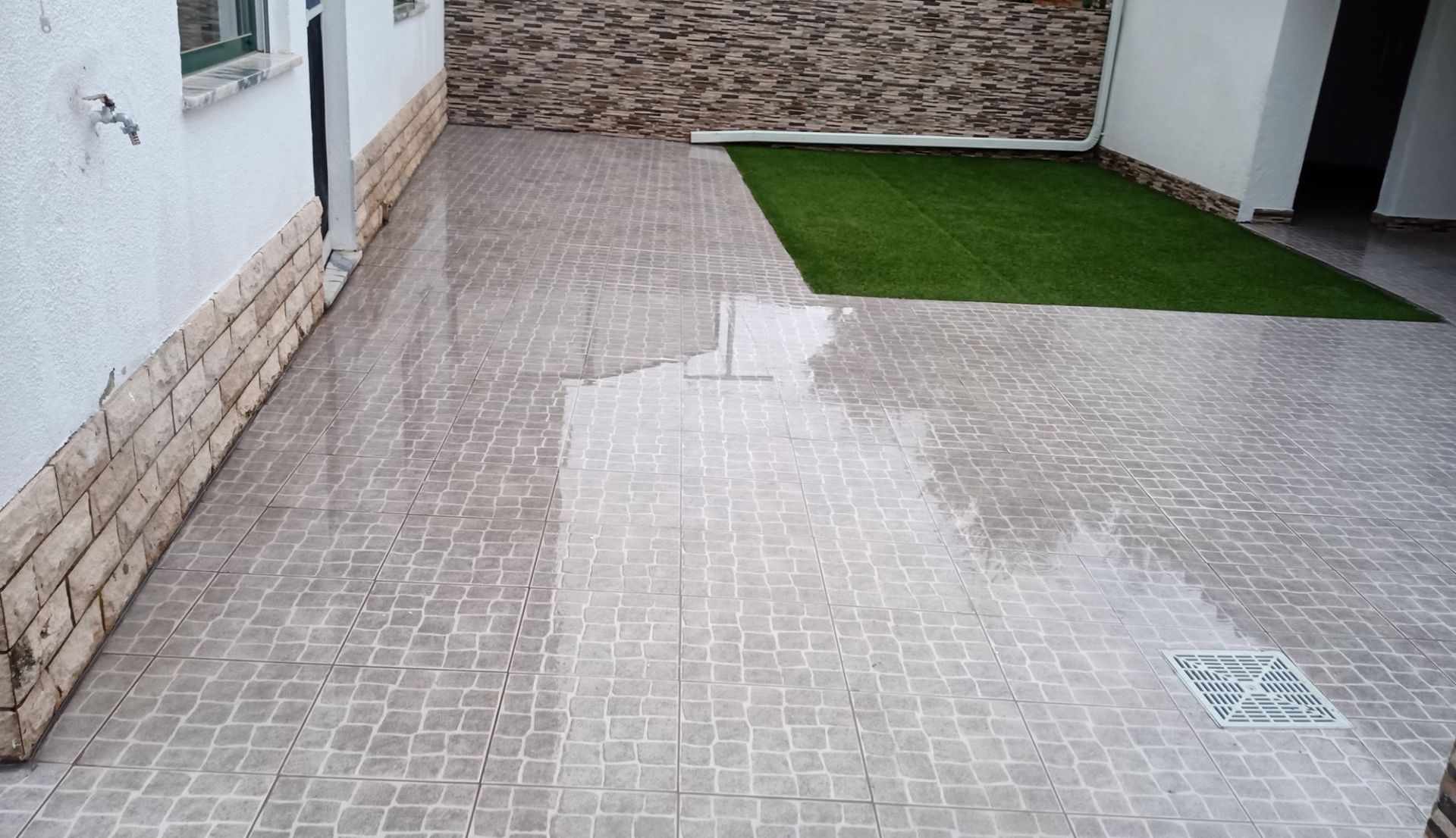 Pátio molhado com piso de azulejo, uma pequena área de grama artificial e uma parede de tijolos como detalhe.