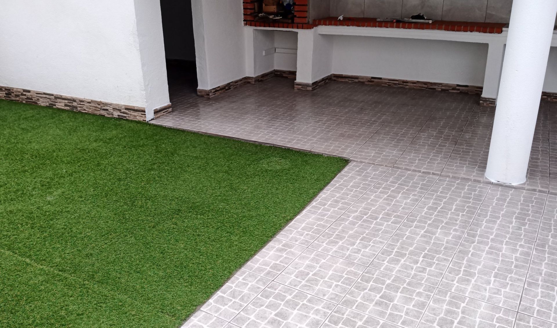 Espaço exterior com relva verde, piso de azulejo, pilares brancos e churrasqueira de tijolo.