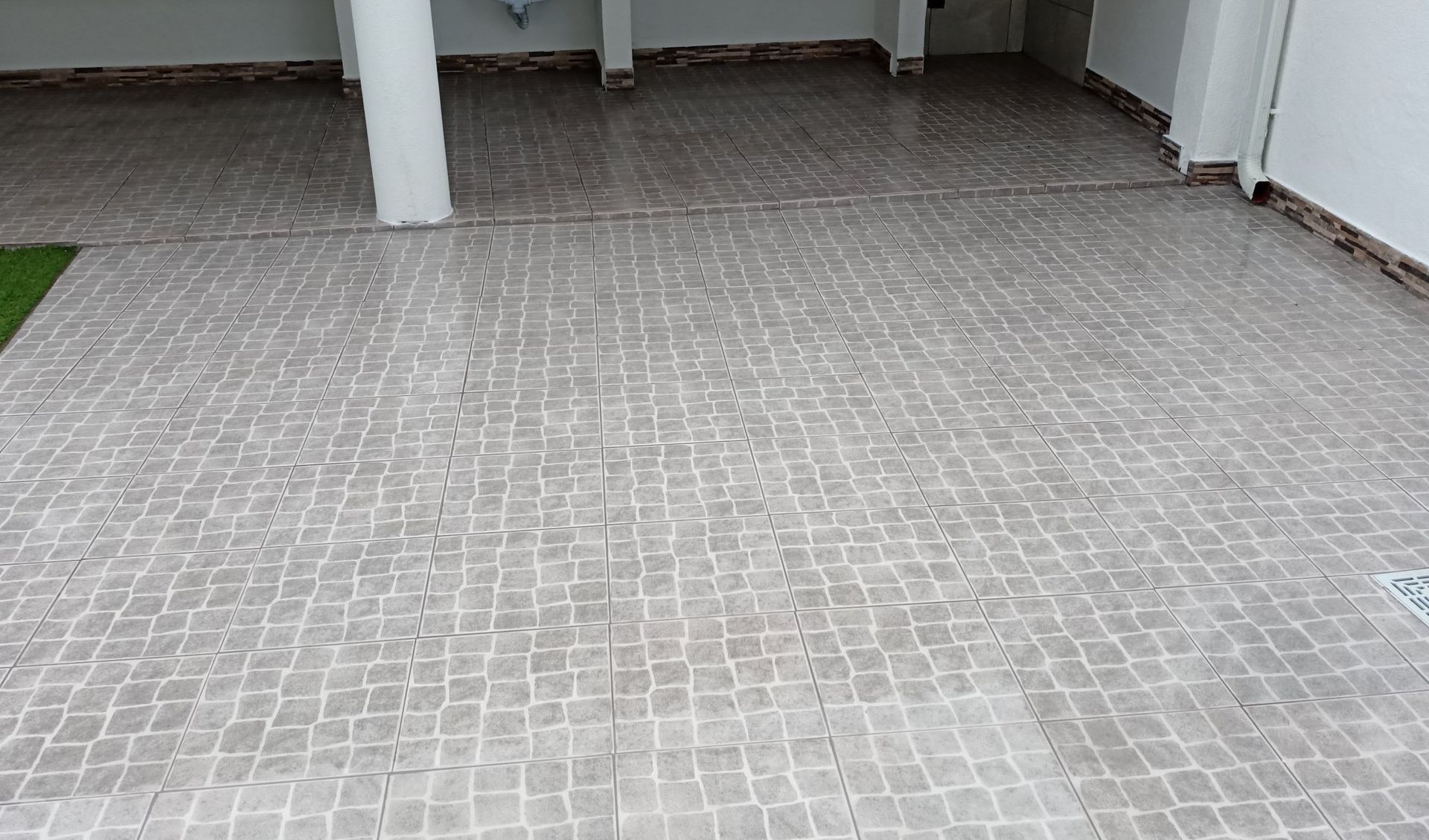 Calçada de paralelepípedos em uma garagem coberta, pedras cinza-claras, pilares e paredes brancas.