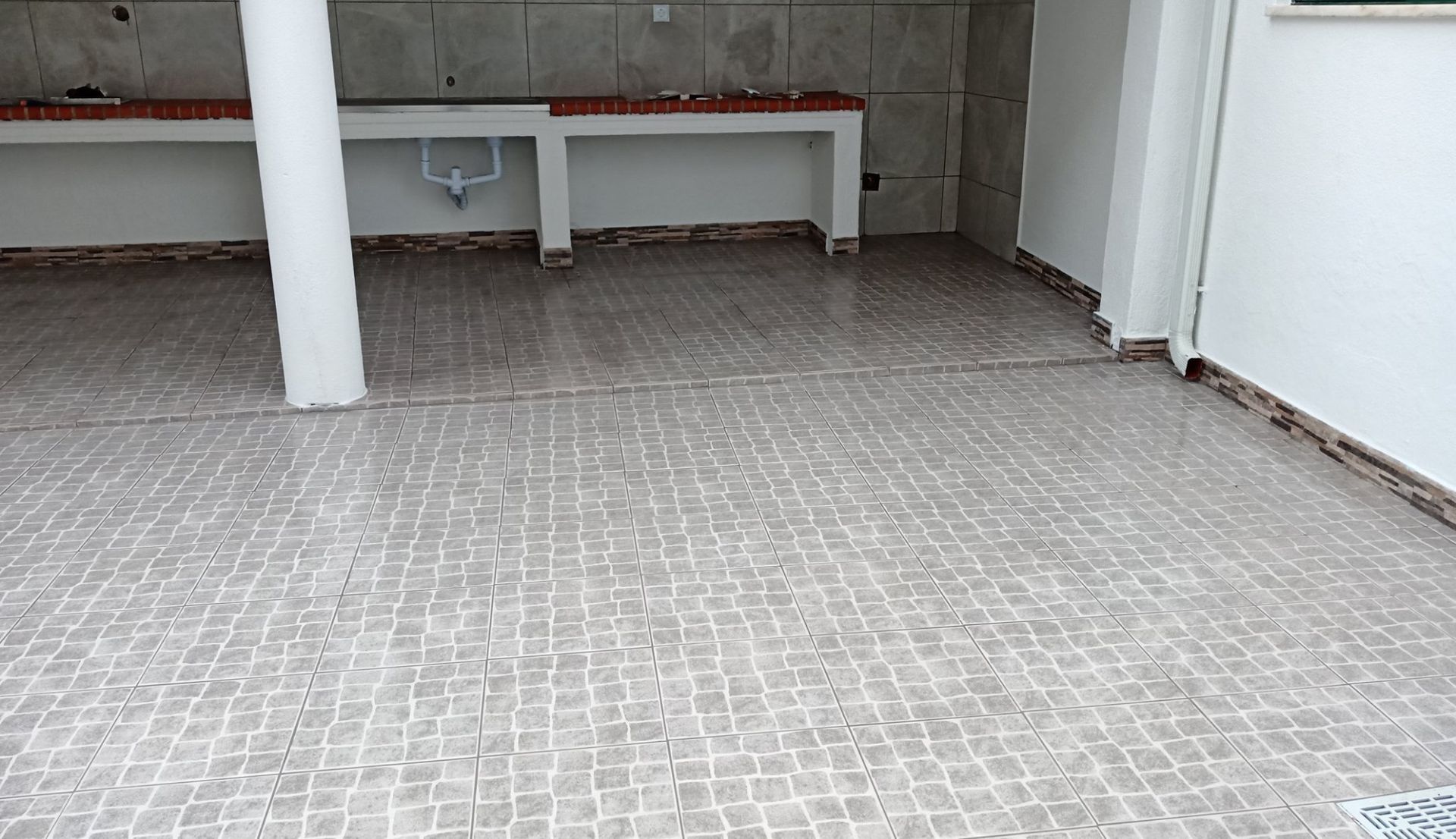 Cozinha externa com piso de azulejo, bancadas brancas e coluna de sustentação.