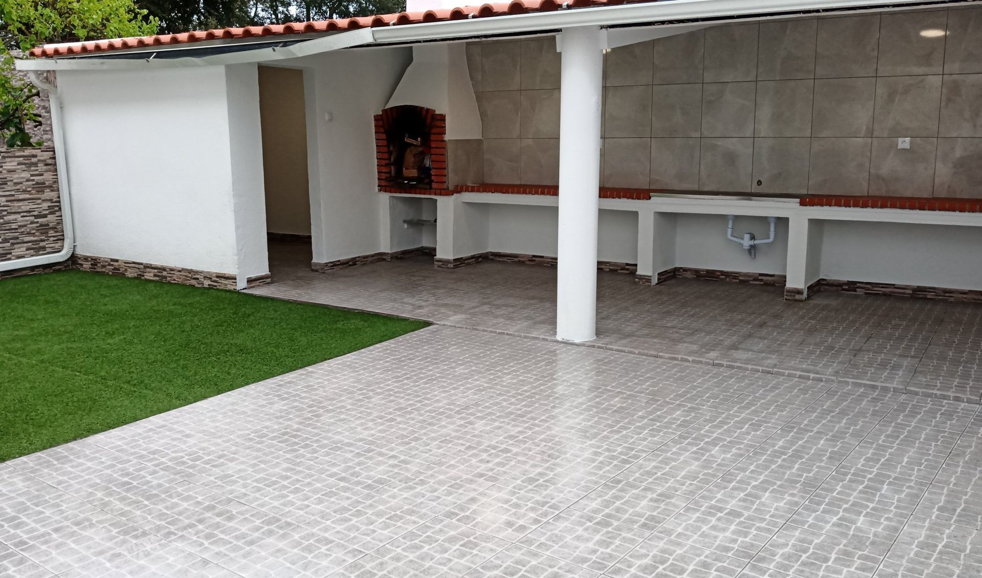 Pátio externo com churrasqueira, bancadas, pilares brancos, piso de azulejo e gramado verde.