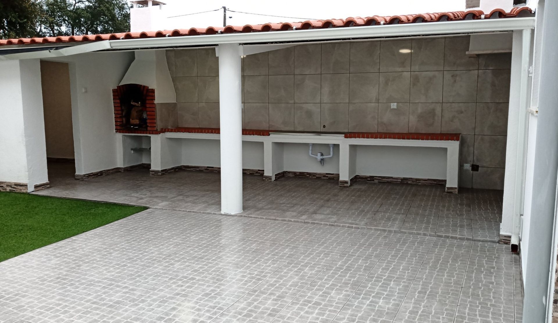 Cozinha externa coberta com piso de azulejo, bancada, churrasqueira e fachada de tijolos.