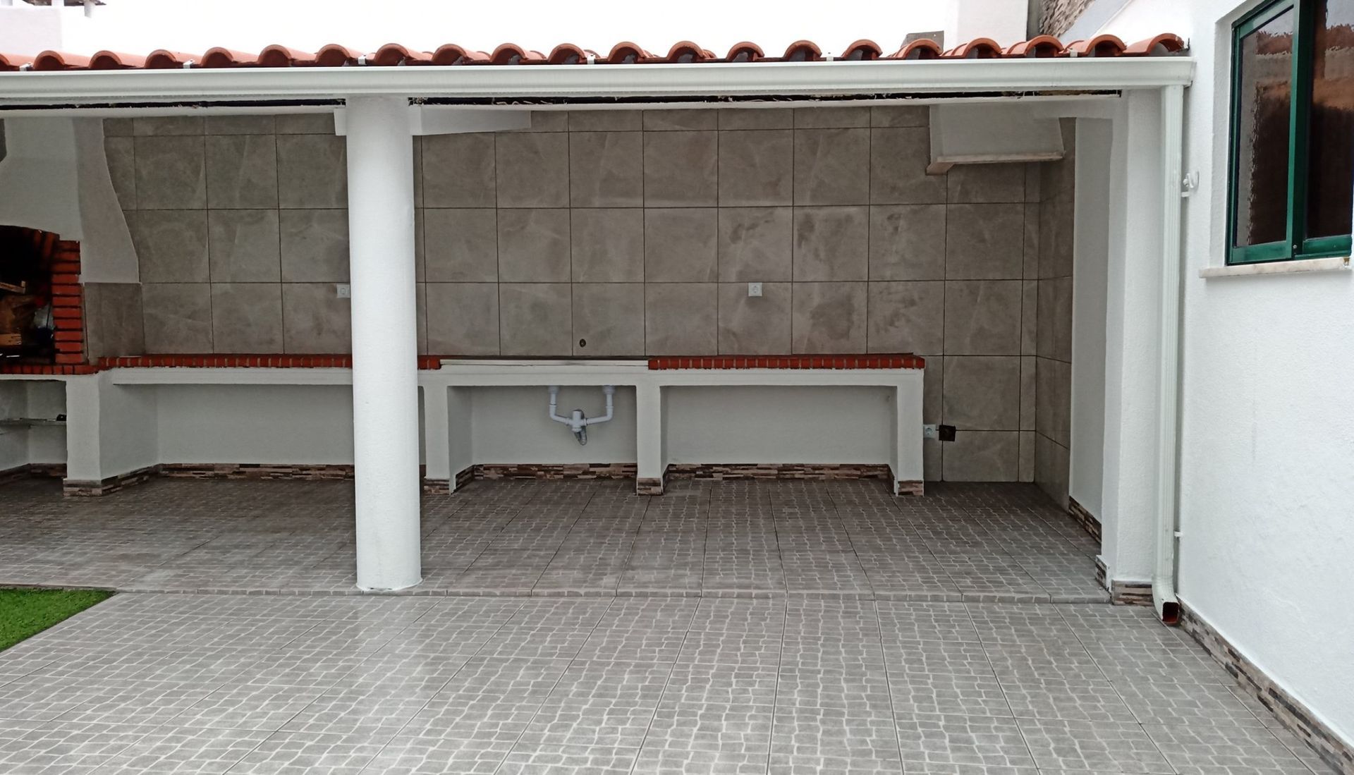 Cozinha externa coberta com churrasqueira e balcão embutidos, colunas brancas, piso e paredes revestidos com azulejos.