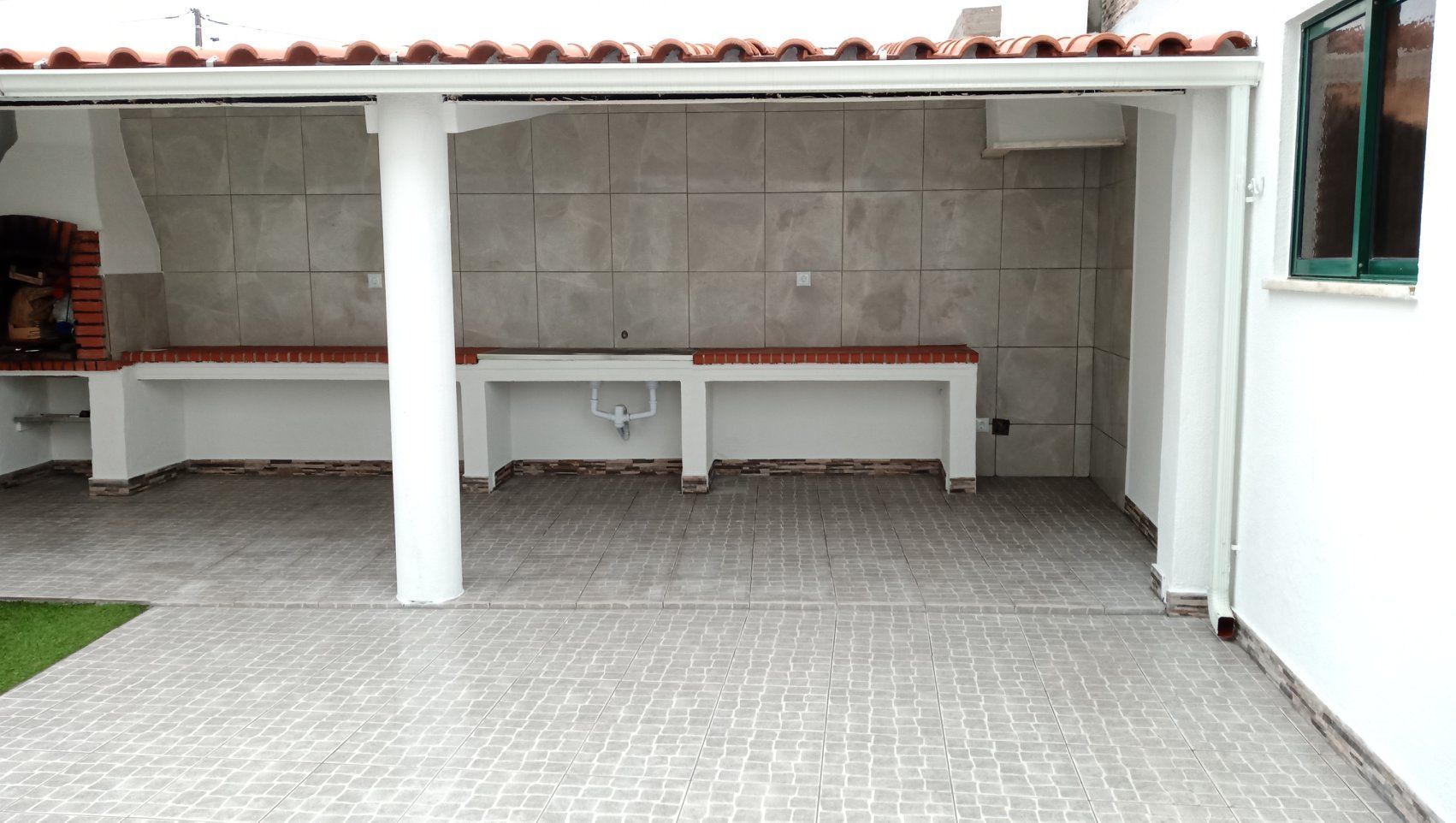 Pátio externo com churrasqueira de tijolos, bancadas de azulejo e colunas brancas.