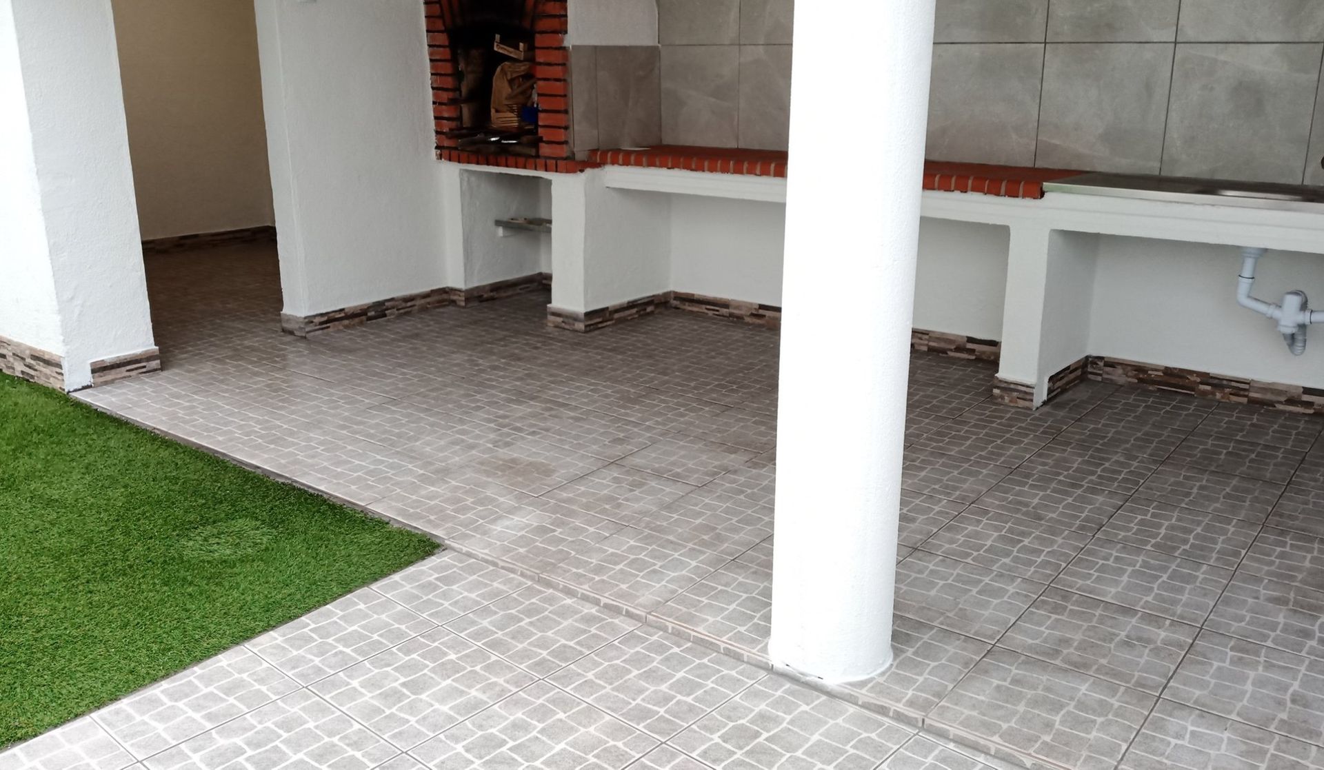 Pátio externo coberto com piso de azulejo, churrasqueira embutida de tijolos e colunas de sustentação brancas.