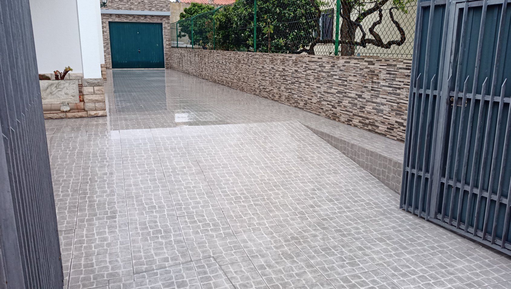 A entrada de veículos é pavimentada com blocos cinza-claros, delimitada por um muro de pedra e um portão cinza fechado.