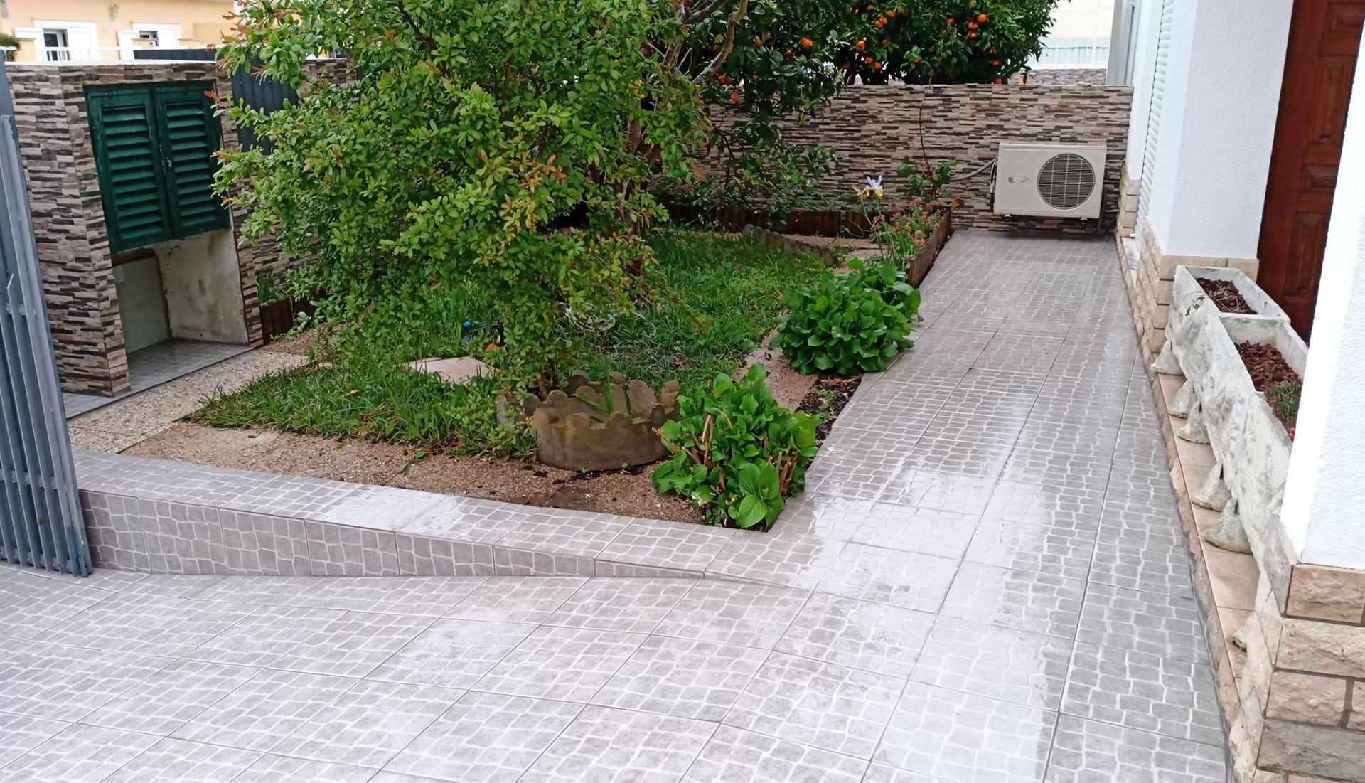 Um pátio com um pequeno jardim, caminho pavimentado e um edifício com persiana fechada e unidade de ar condicionado.