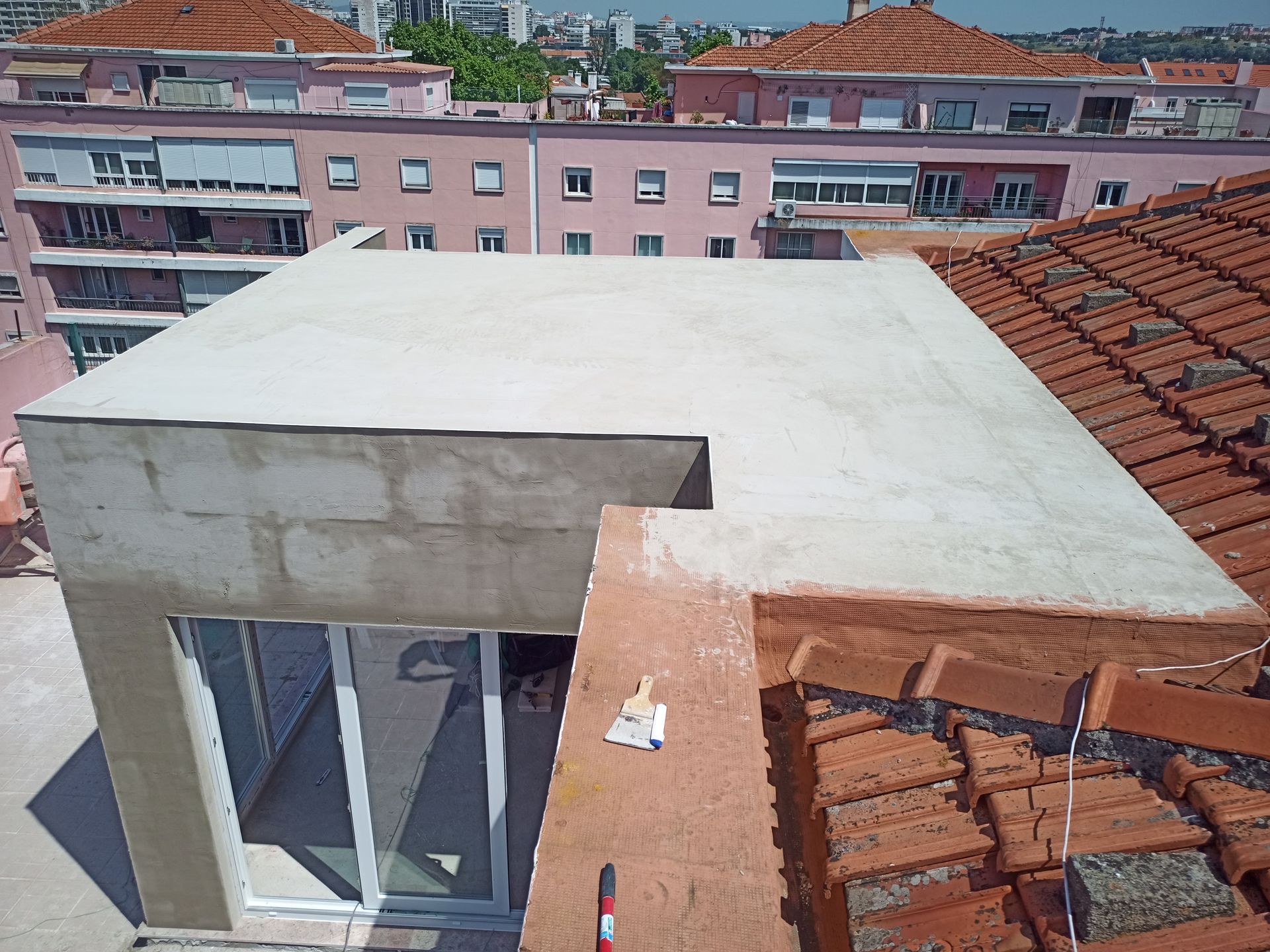 Ampliação do telhado em construção; telhado plano branco com janela, em contraste com o telhado de telhas e os edifícios da cidade.