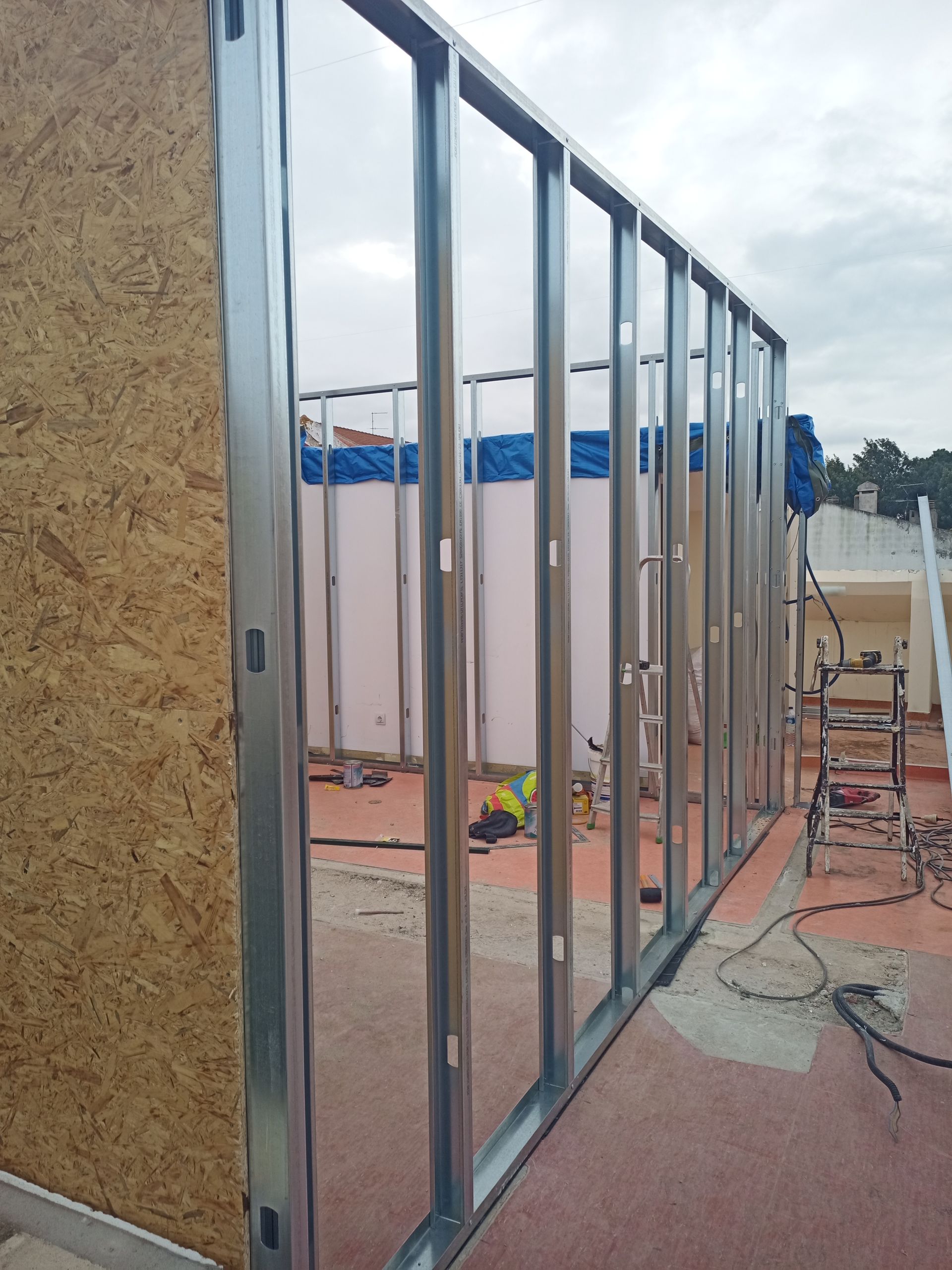 Estrutura de parede com montantes metálicos parcialmente revestida com painéis de madeira; canteiro de obras ao ar livre.