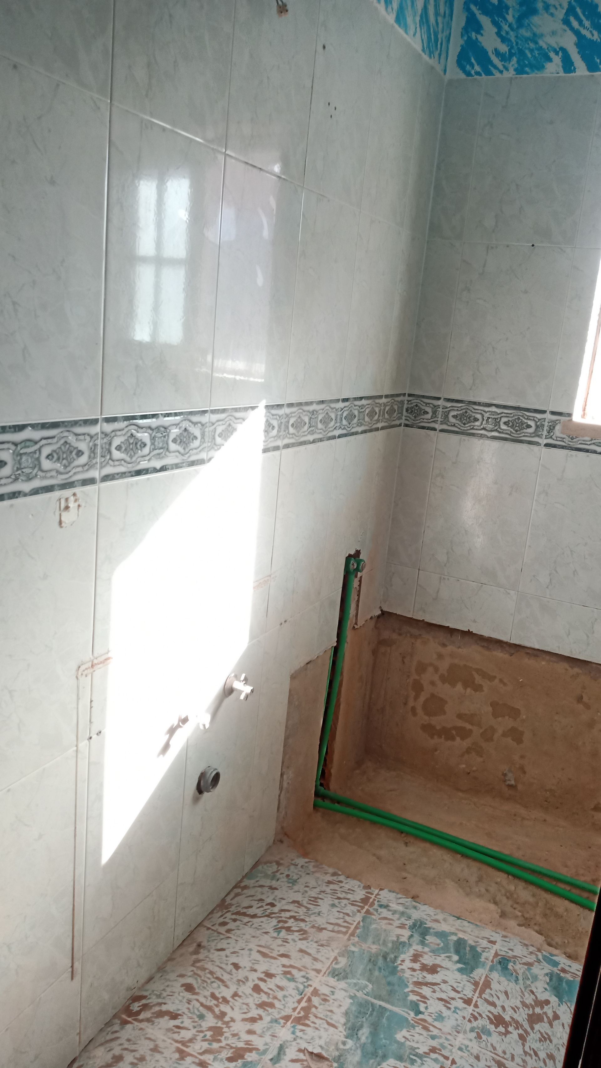 Interior de banheiro com paredes revestidas de azulejos, parte inferior inacabada, encanamento aparente e janela.