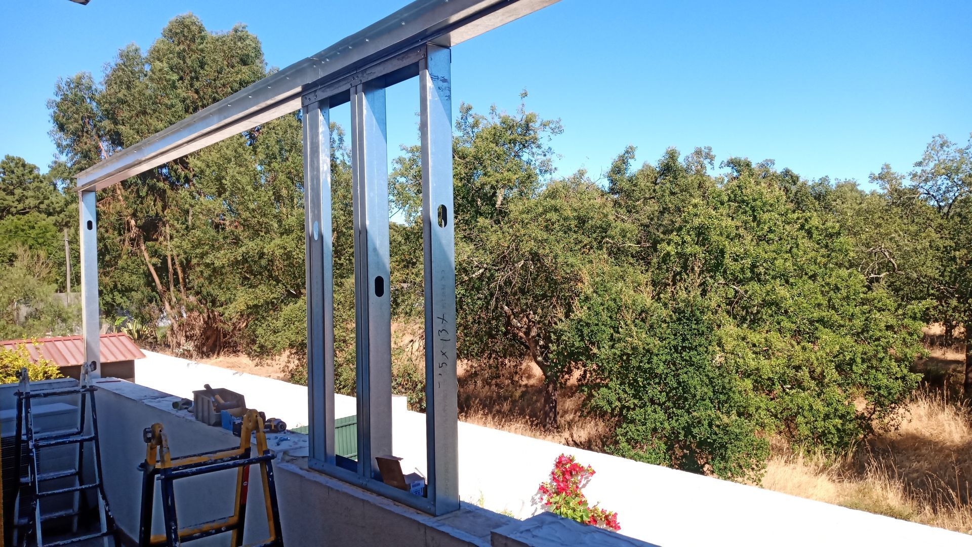 Estrutura metálica de uma porta de correr de vidro sendo instalada em uma varanda, com vista para as árvores e o céu.