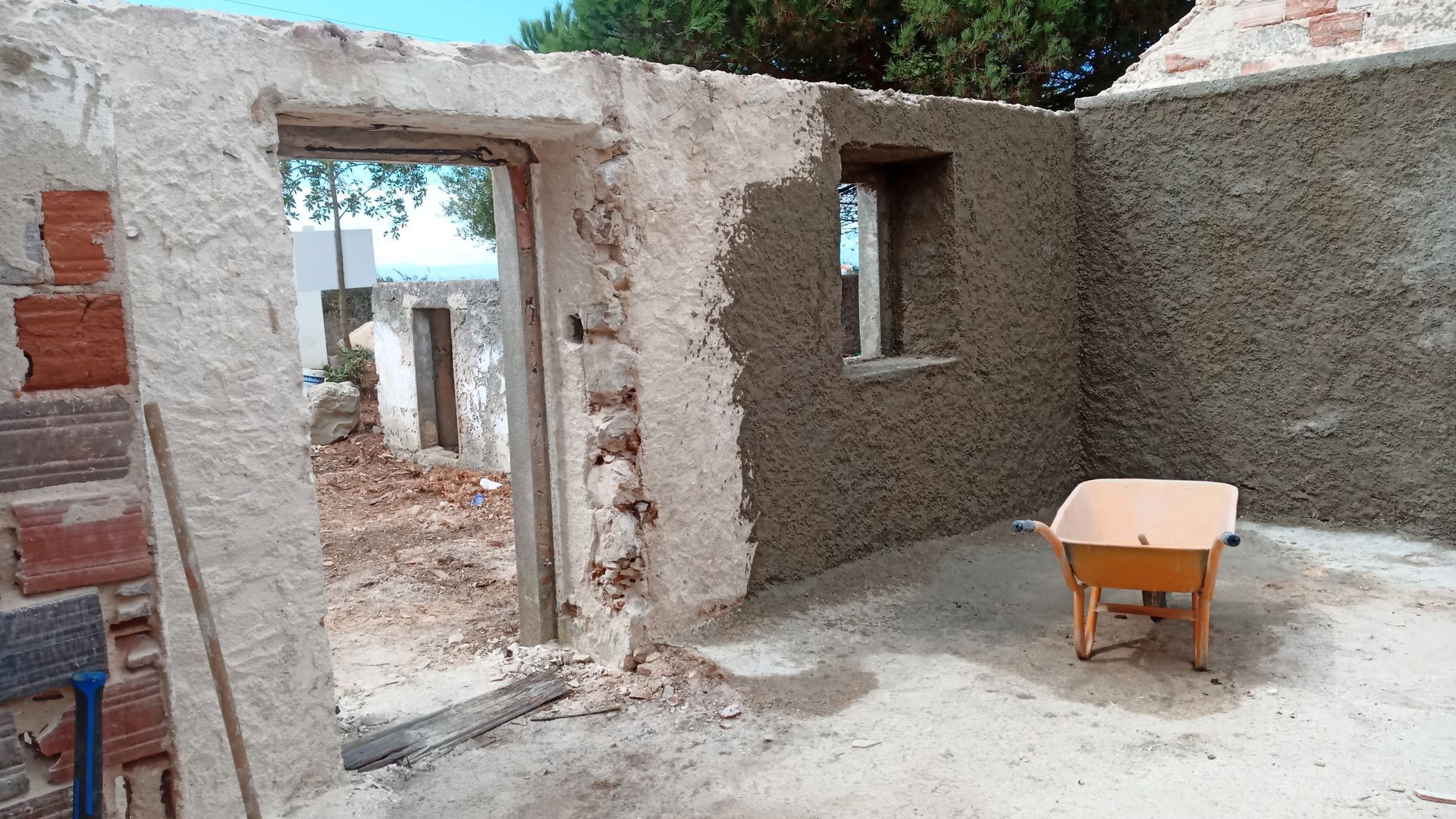 Interior de um edifício em construção, mostrando uma porta, uma janela e paredes parcialmente rebocadas. Um carrinho de mão está dentro do edifício.