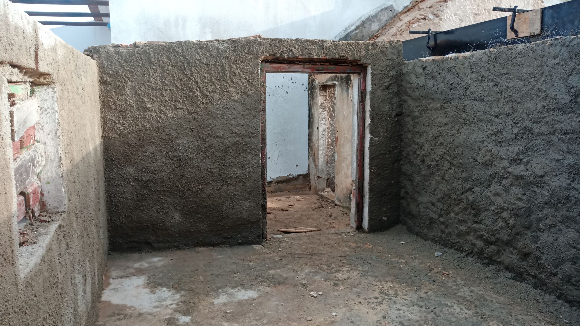 Interior de uma sala com paredes de cimento inacabadas, porta e piso de concreto; aparenta estar em construção.