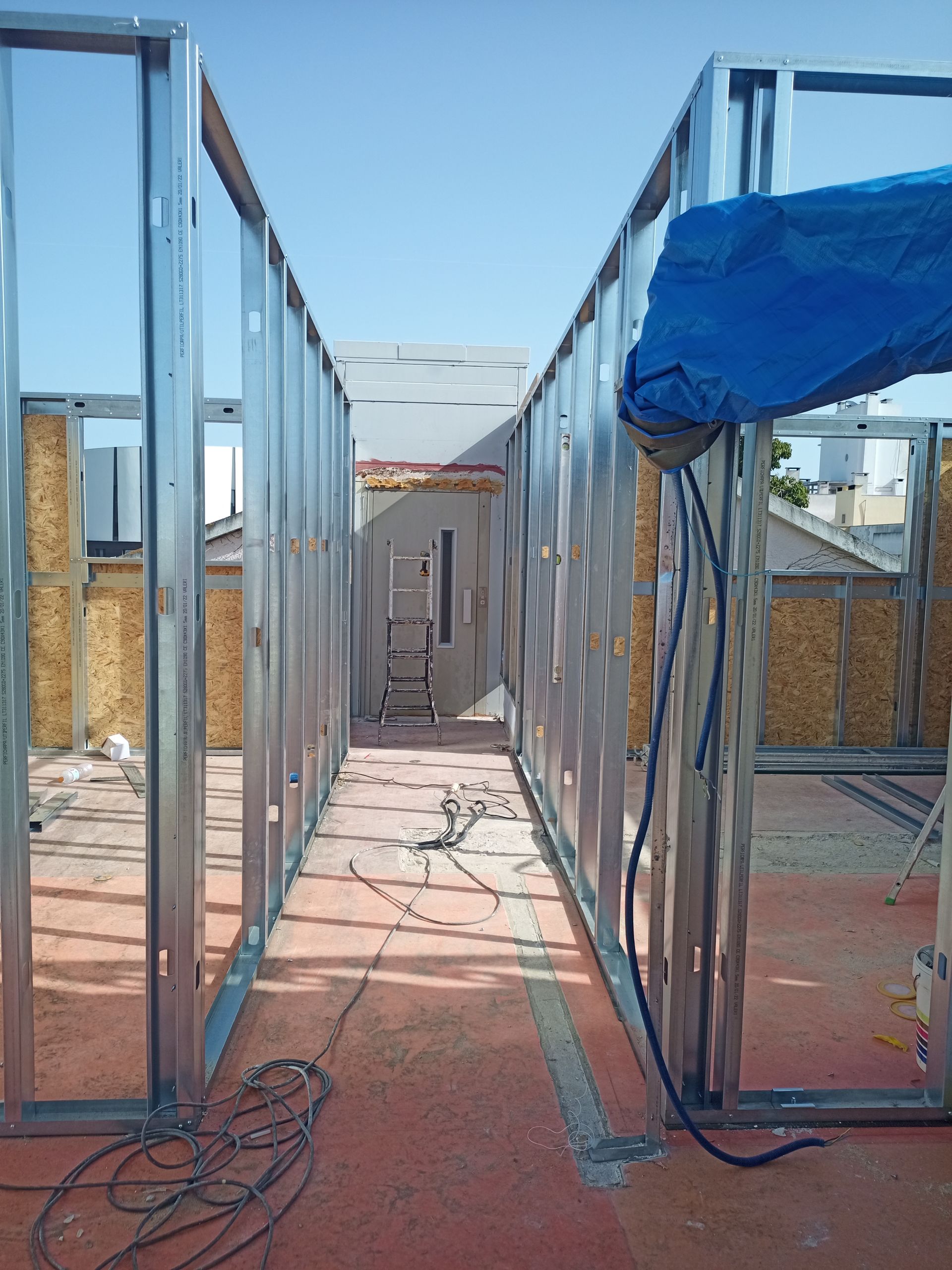 Estrutura metálica das paredes internas; canteiro de obras coberto com uma lona azul em um dia ensolarado.