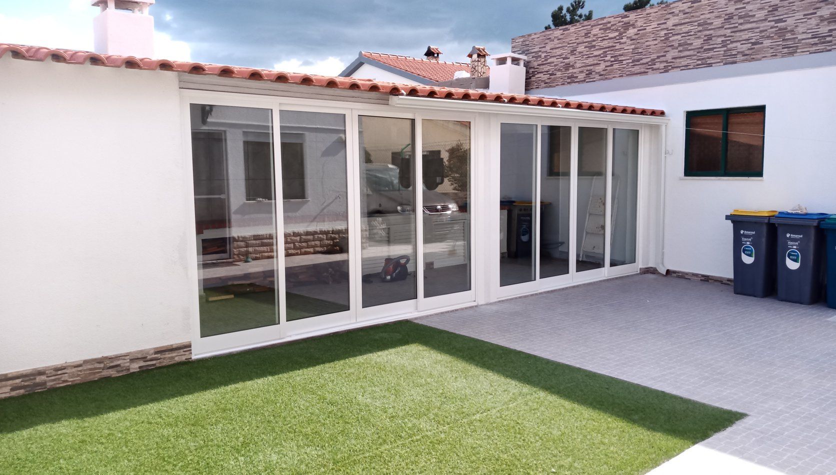 Vista exterior de uma varanda envidraçada com portas de correr de vidro, adjacente a um pequeno gramado. Paredes brancas, telhado de telha.