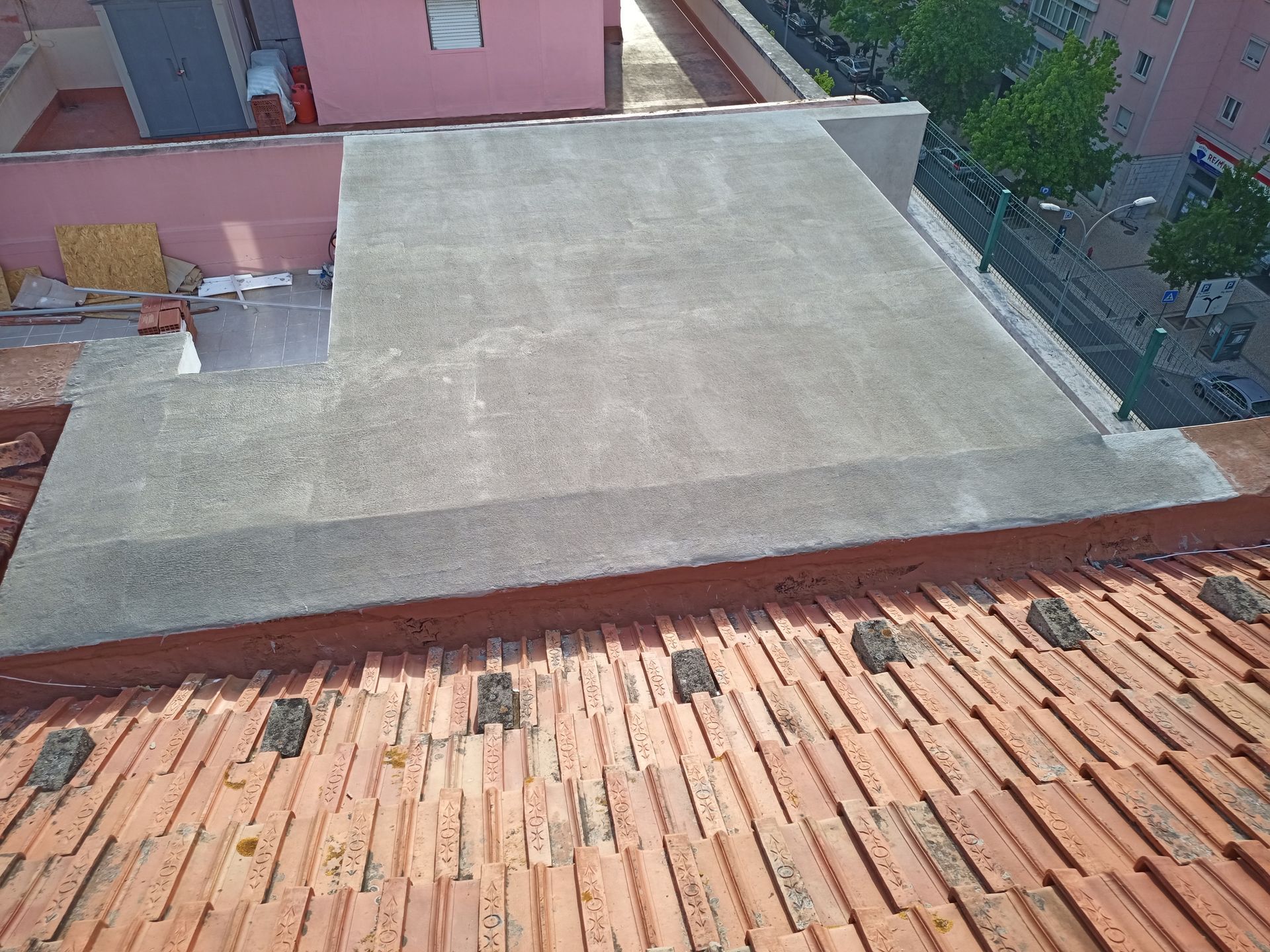 Vista de um telhado com telhas laranja em primeiro plano e uma estrutura de concreto ao centro, com rua e árvores visíveis.