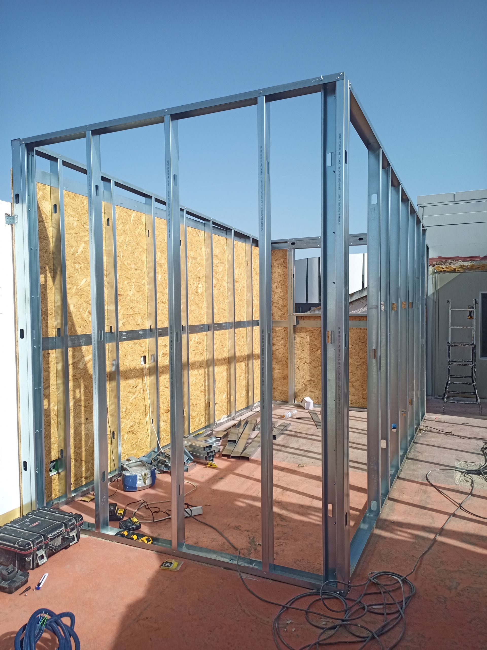 Estrutura metálica de um pequeno edifício em construção, com painéis de madeira parcialmente instalados. Céu azul ao ar livre.
