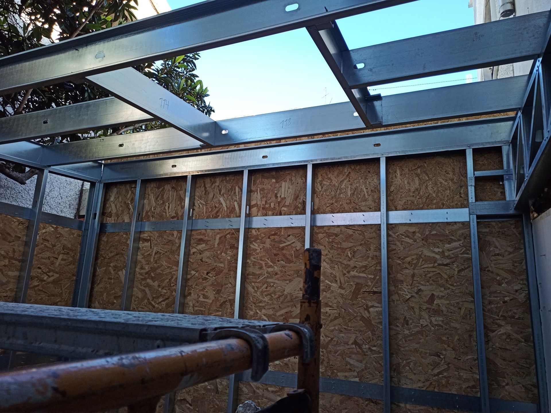 Construção com estrutura metálica, paredes em placa OSB e céu visível através de um teto aberto.