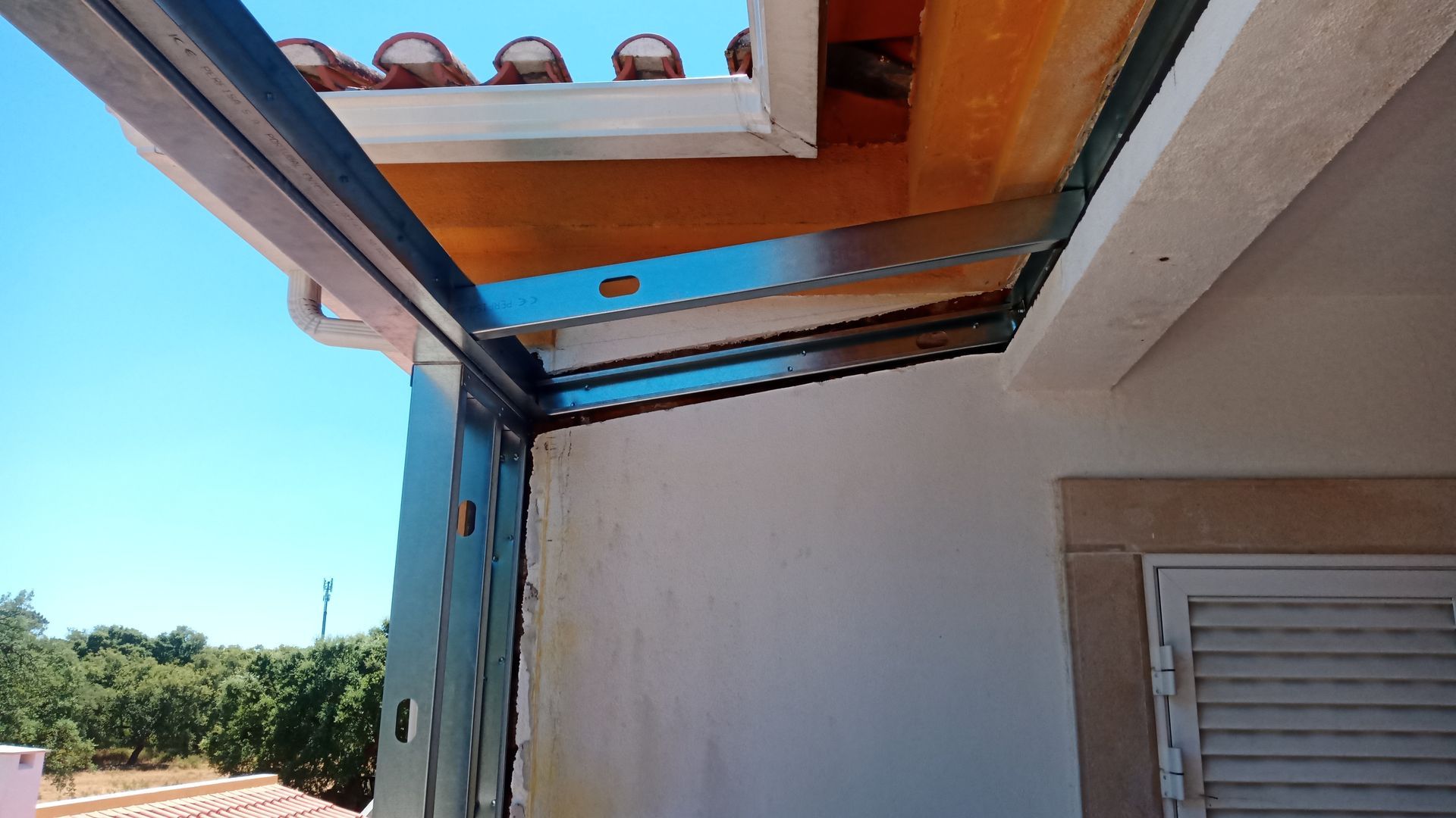 Caixilharia metálica para janela ou porta, instalada num edifício com telhas de terracota e paredes brancas, contra um céu azul.