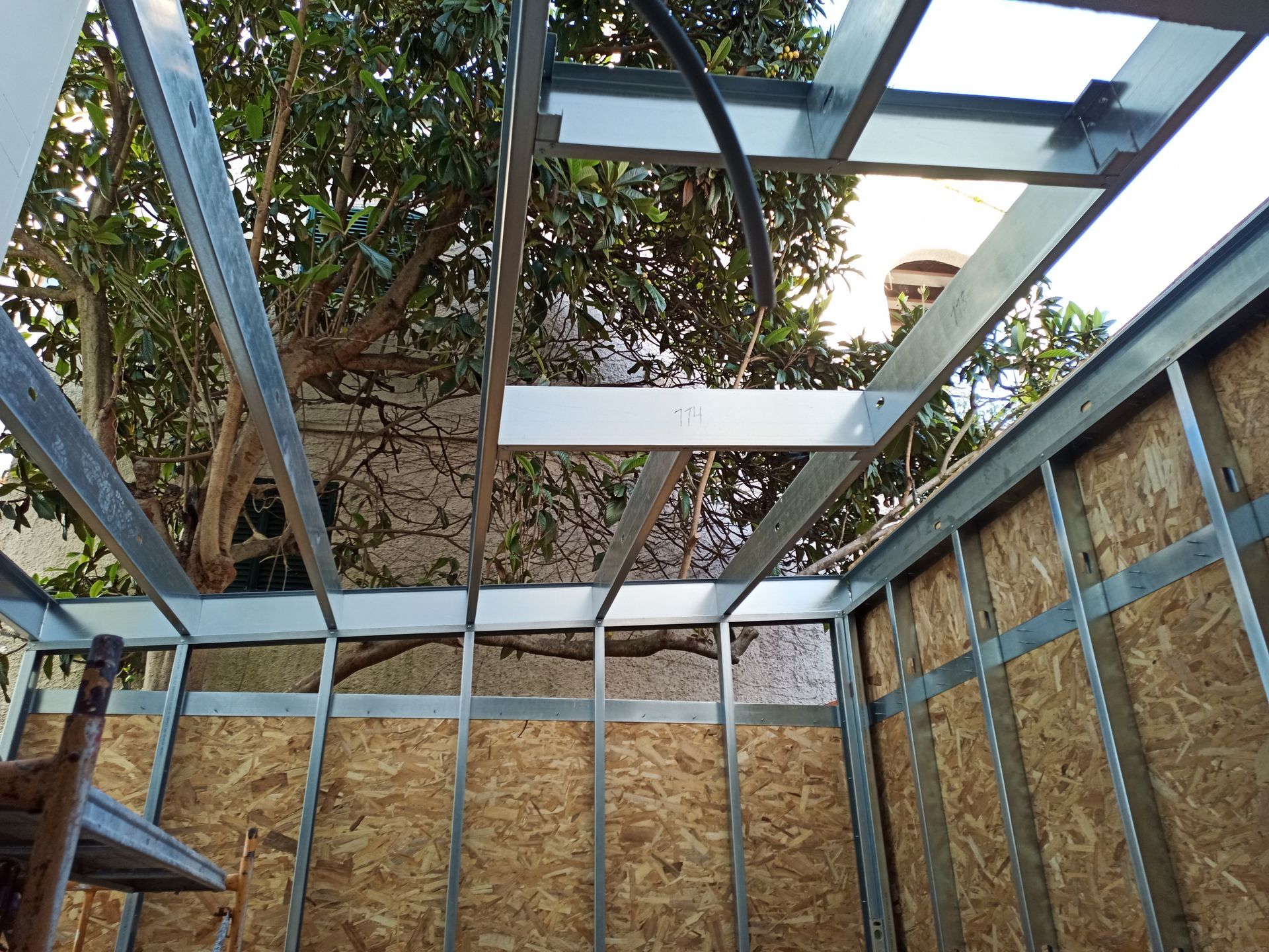 Estrutura metálica com paredes em OSB, teto parcialmente aberto, área externa com árvore.