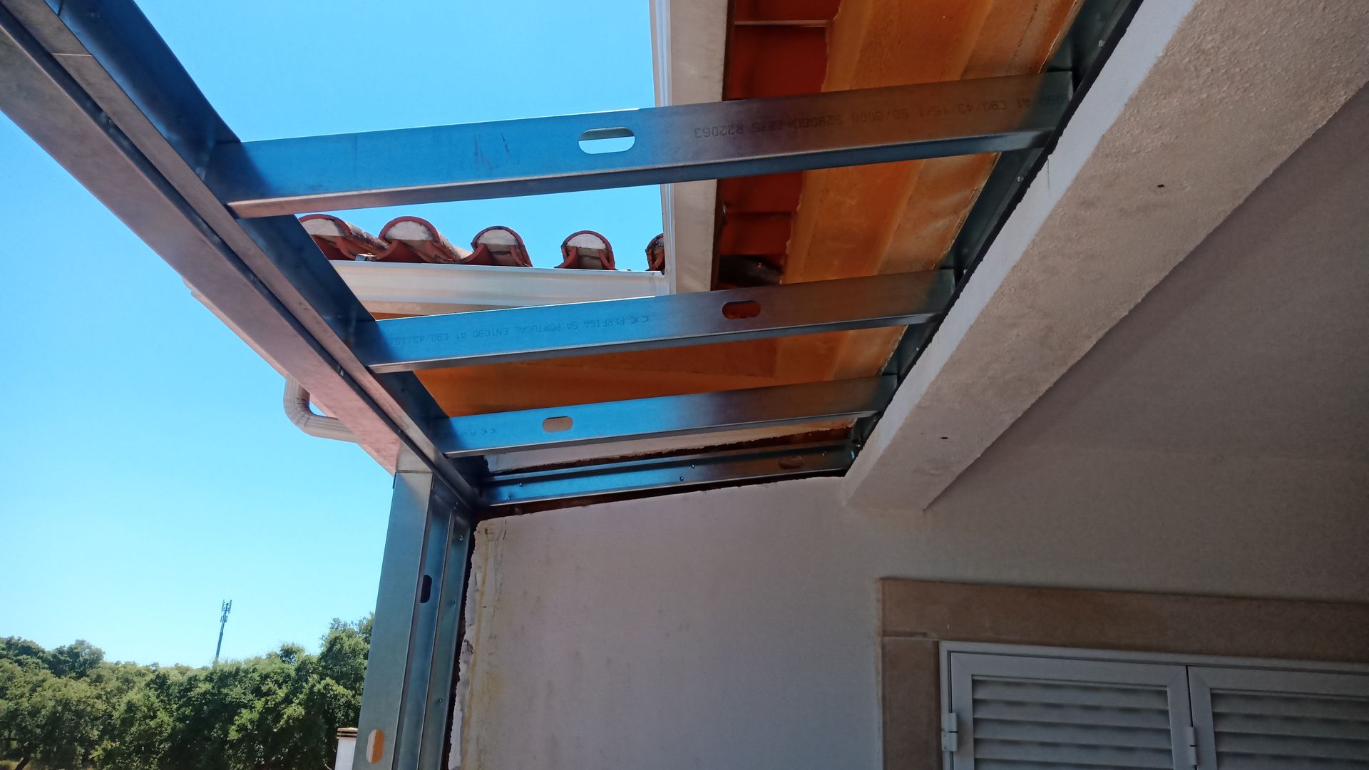 Estrutura metálica com suportes de madeira sob um telhado de telhas, contra um céu azul.