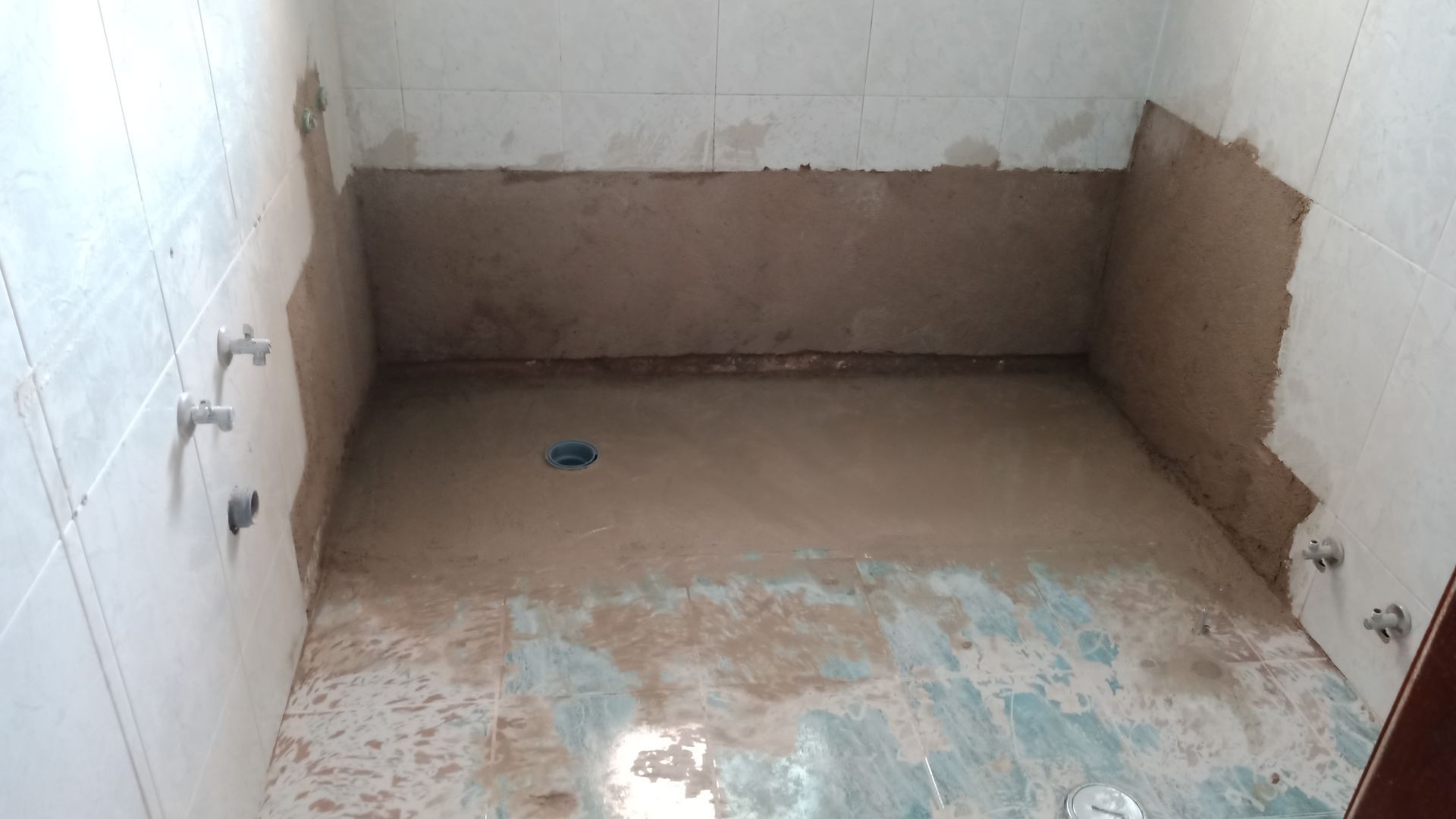 Banheiro em construção. Piso e paredes de cimento marrom, encanamento aparente, azulejos parcialmente instalados.