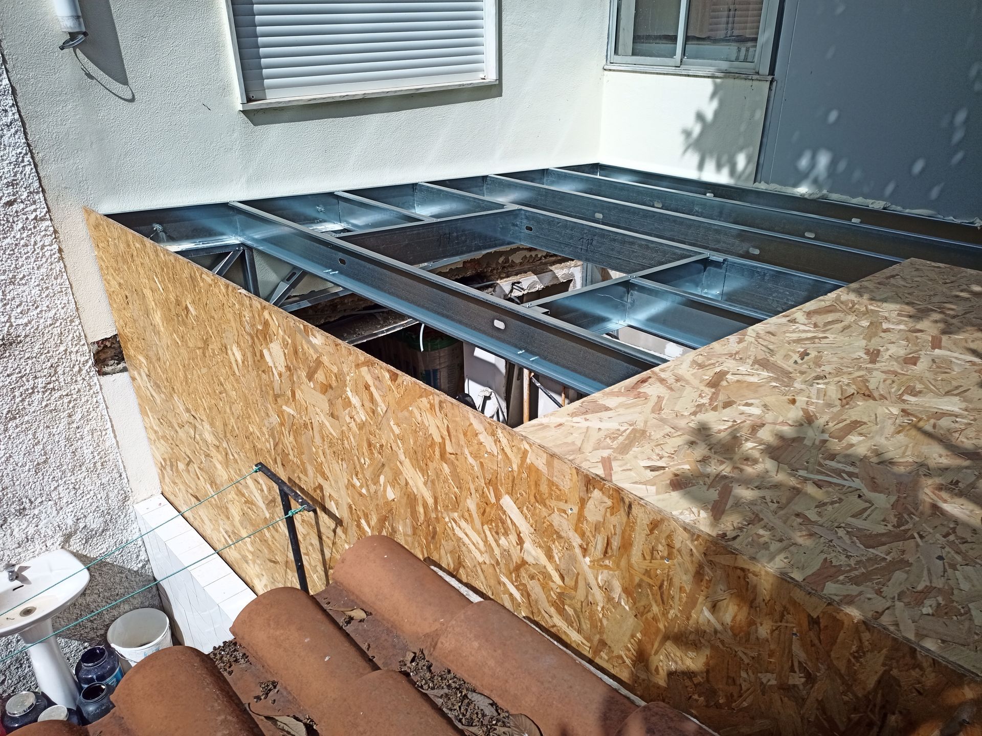 Construção de um novo deck com estrutura metálica e placas de OSB, ao lado de um edifício com paredes brancas.