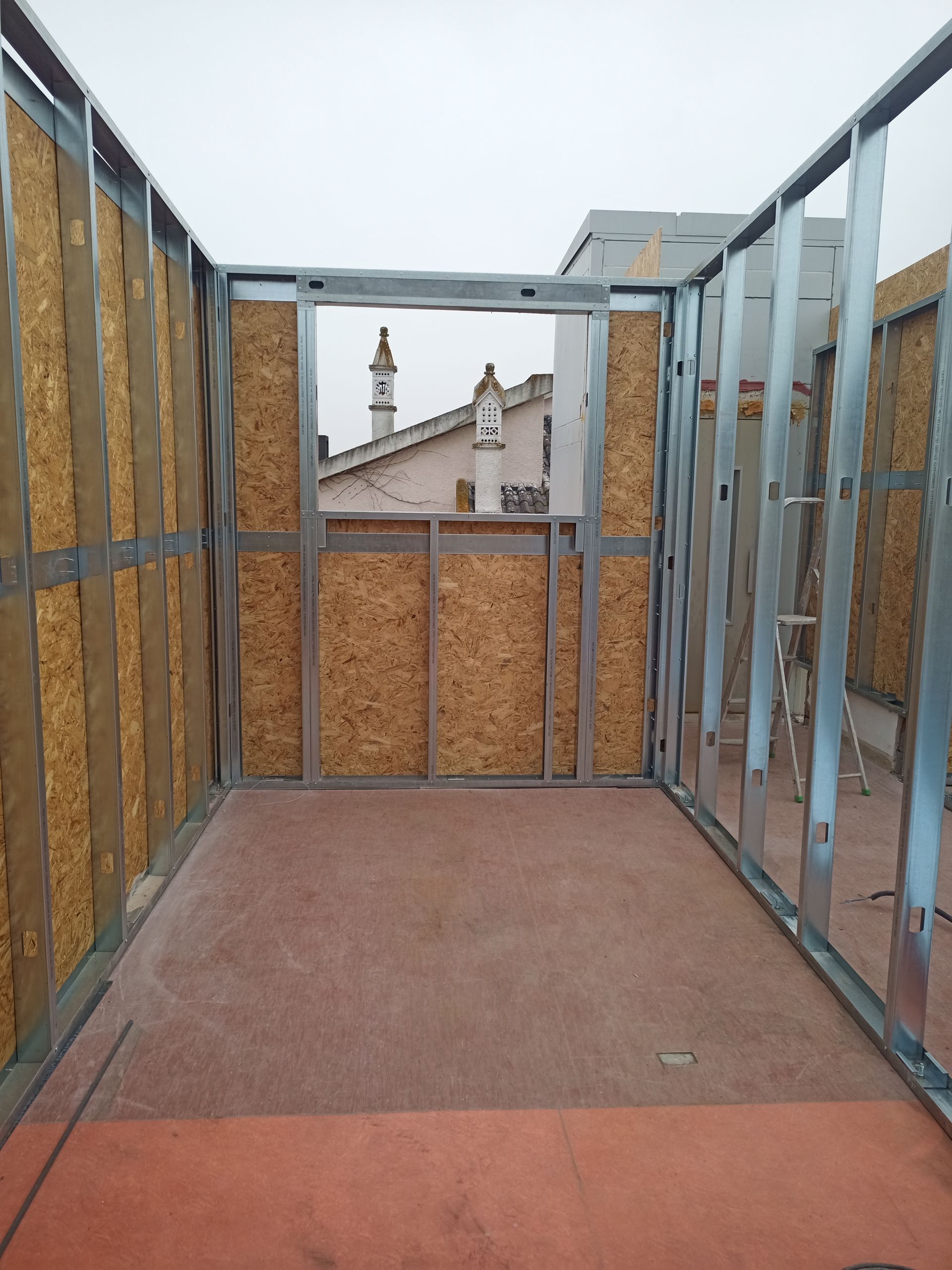 Estrutura interna de um cômodo, com vigas metálicas e painéis de partículas. Uma janela oferece vista para o telhado.