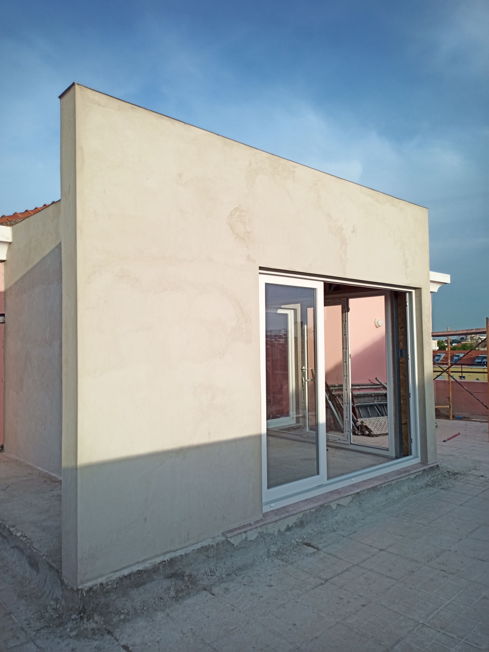 Exterior de um edifício em construção, com uma parede bege e uma porta de correr de vidro, contra um céu azul.