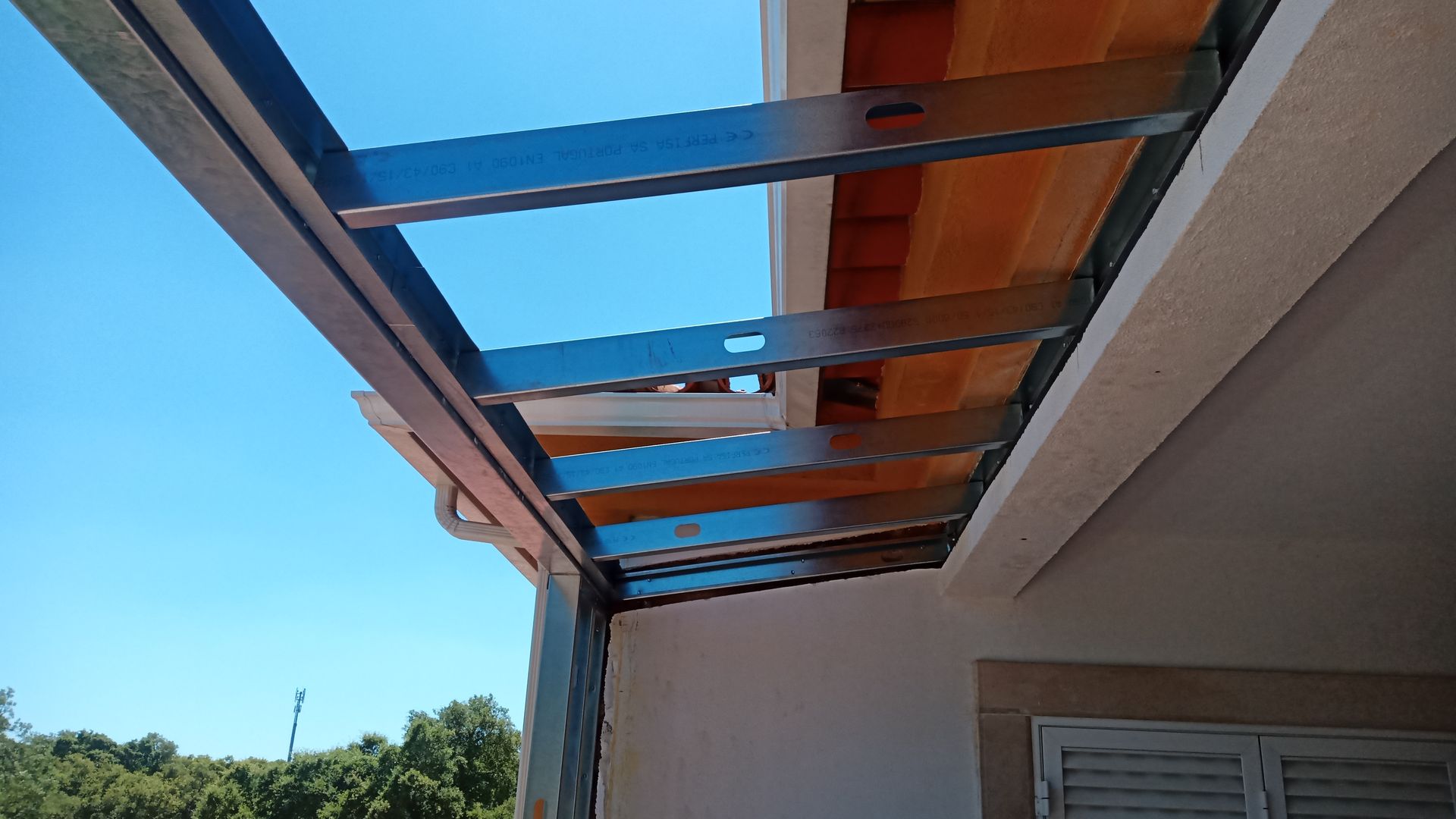Vigas metálicas formando uma pérgola na fachada de um edifício, contra um céu azul brilhante.