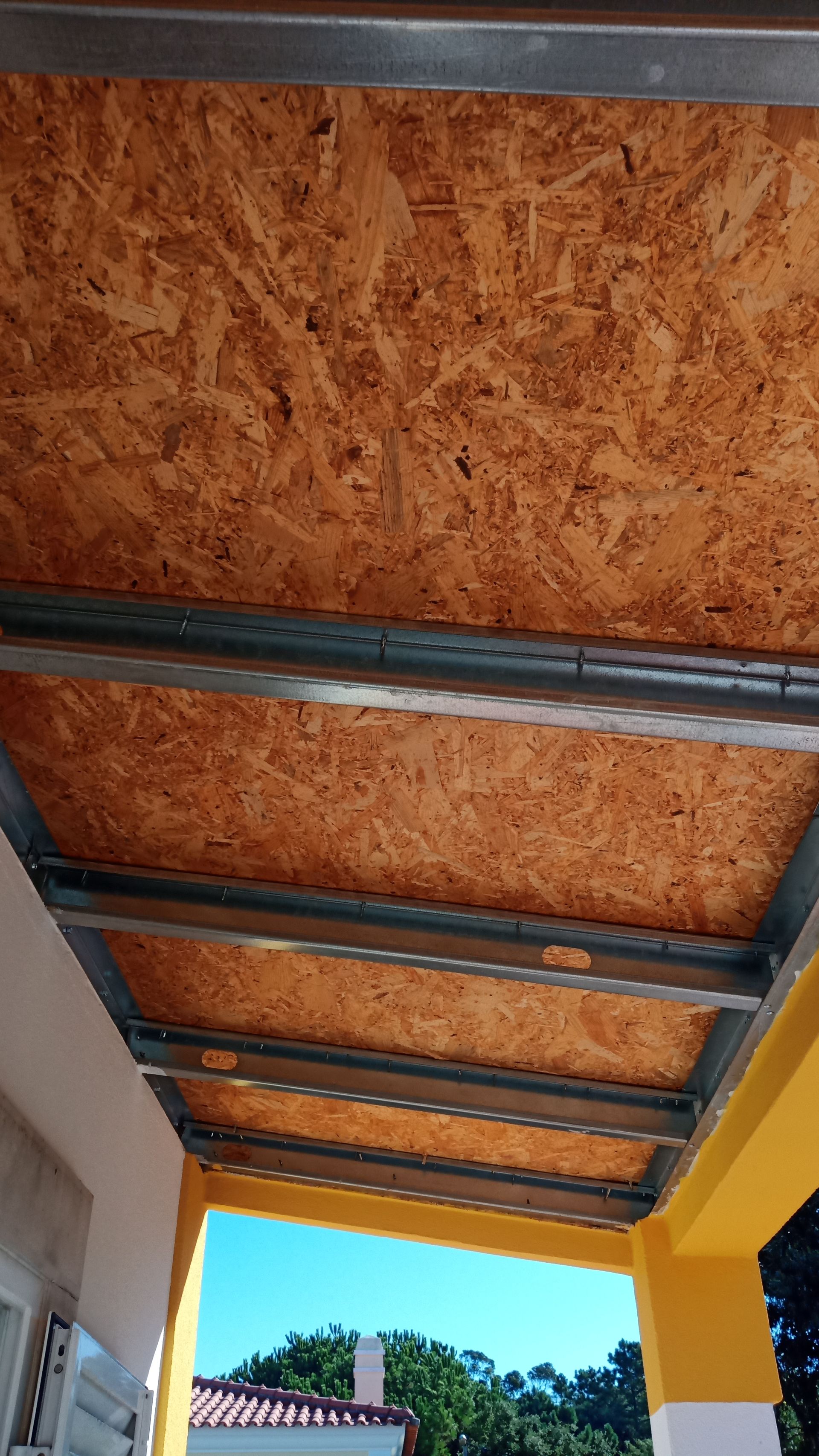 Teto com painéis de madeira e estrutura metálica, sob um céu azul.