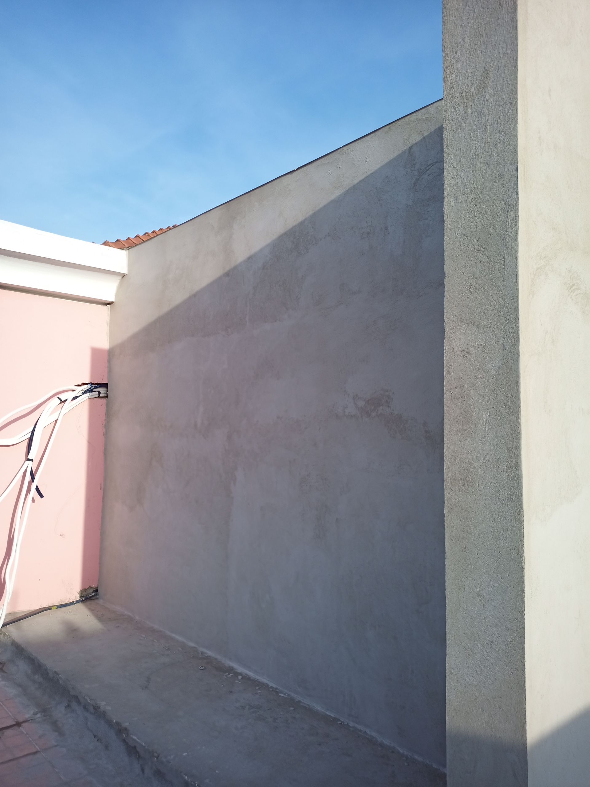 Parede de estuque cinza sob a luz do sol, ao lado de uma parede rosa e uma coluna branca. Céu azul claro.