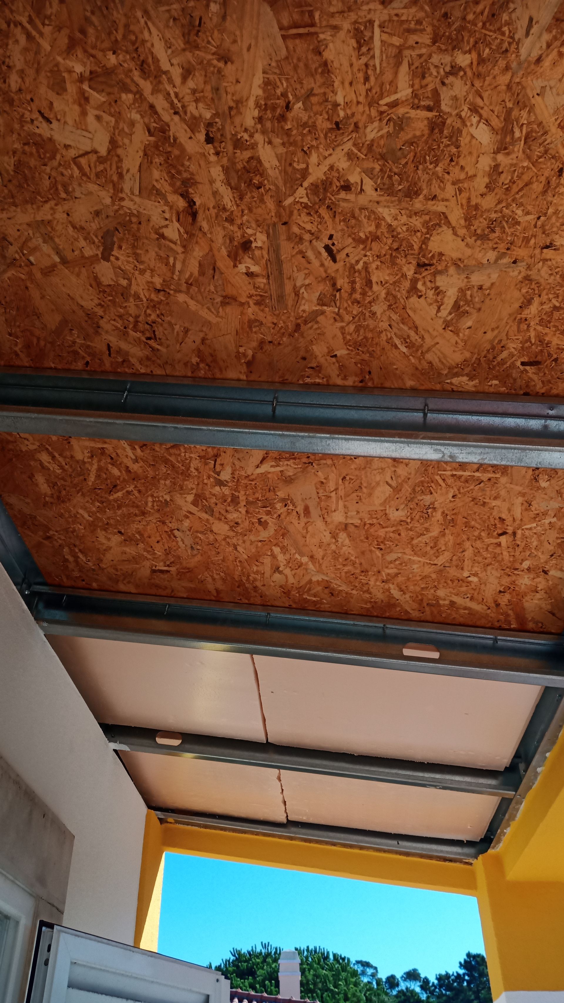 Teto de OSB sob estrutura metálica; varanda com painéis de teto brancos e parede amarela, céu azul visível.