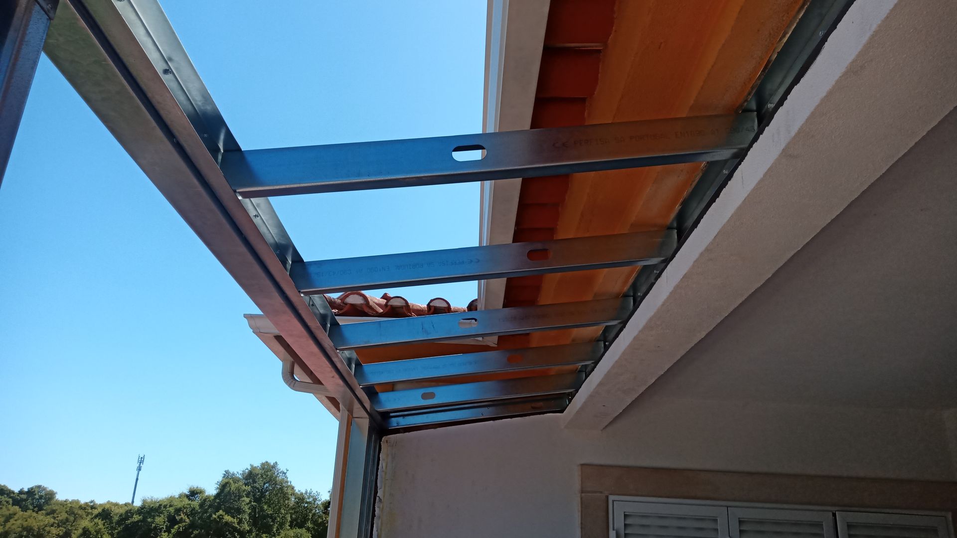 Estrutura metálica para toldo, fixada a um edifício branco com telhado de telha vermelha, contra um céu azul.