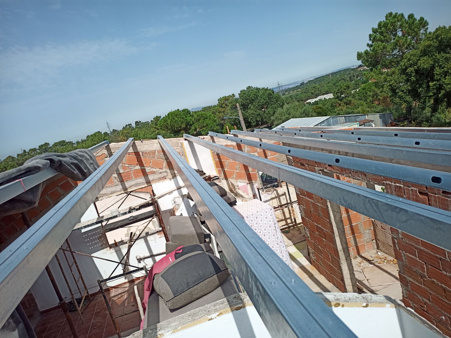 Construção da estrutura do telhado de um prédio de tijolos com uma pessoa trabalhando. Dia ensolarado com árvores ao fundo.