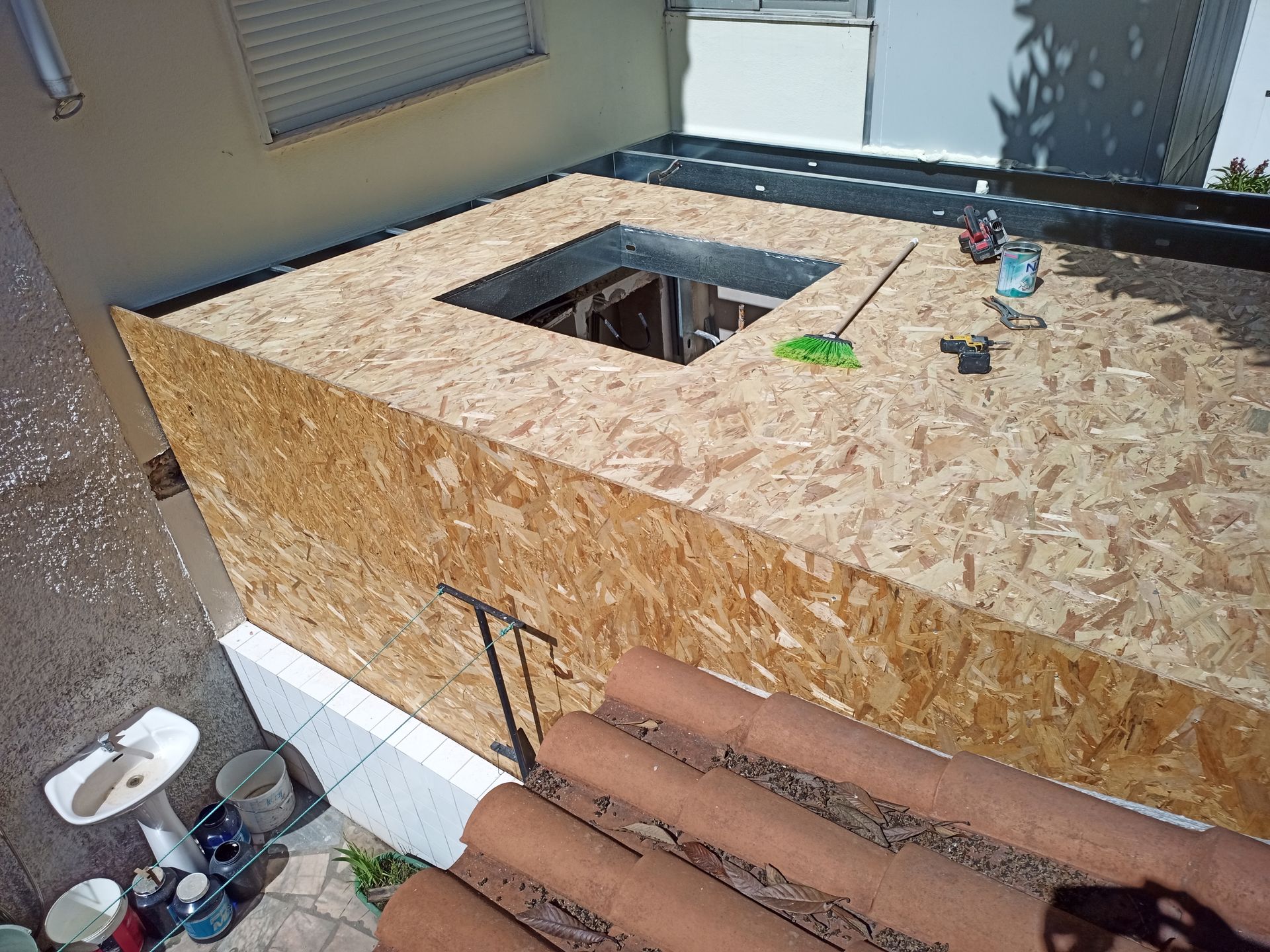 Construção de uma estrutura de cobertura com uma abertura central. Placas de OSB marrom, ferramentas e telhas de barro visíveis.