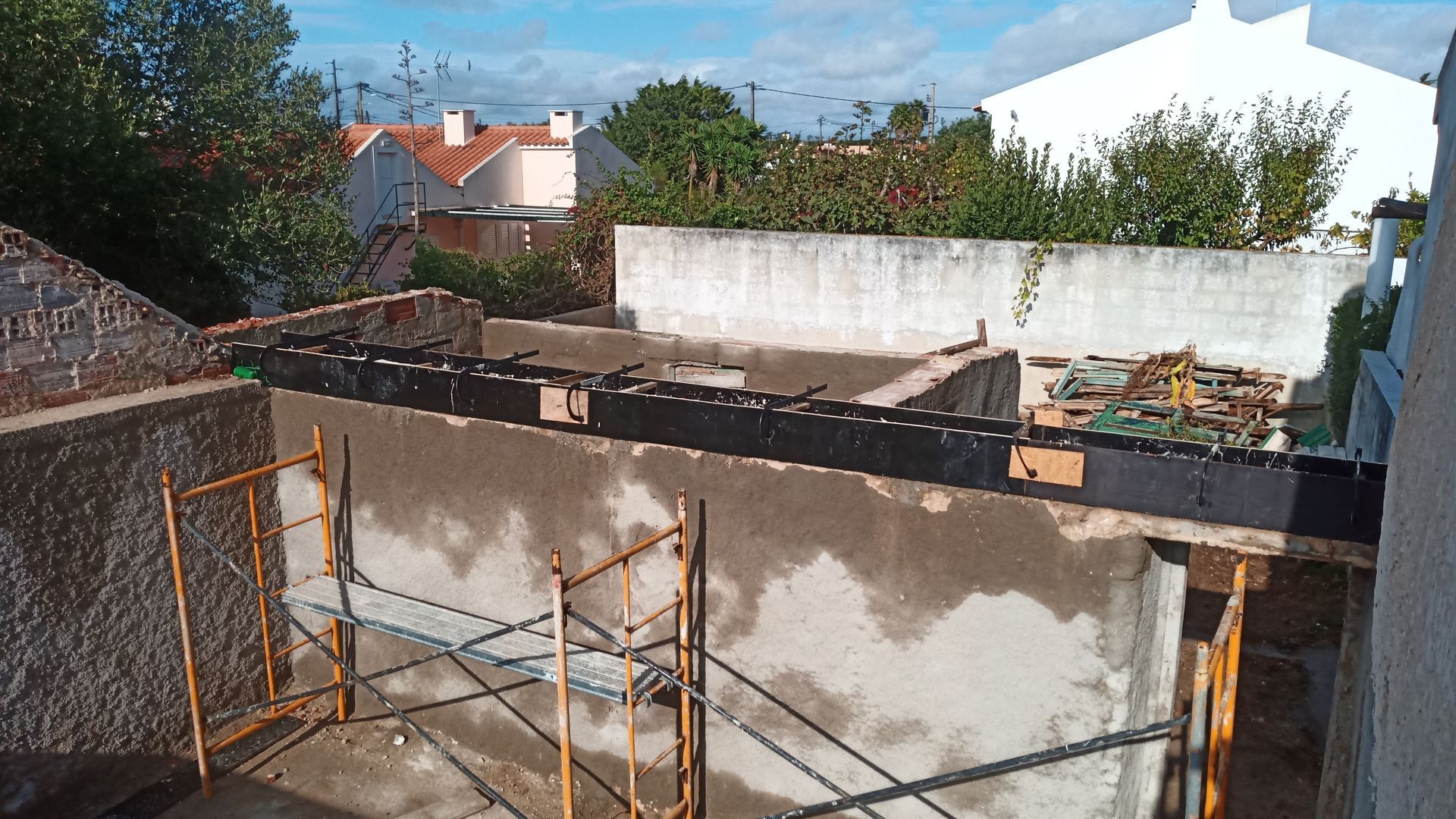 Canteiro de obras: paredes de concreto, andaimes, vigas pretas e prédios residenciais ao fundo.