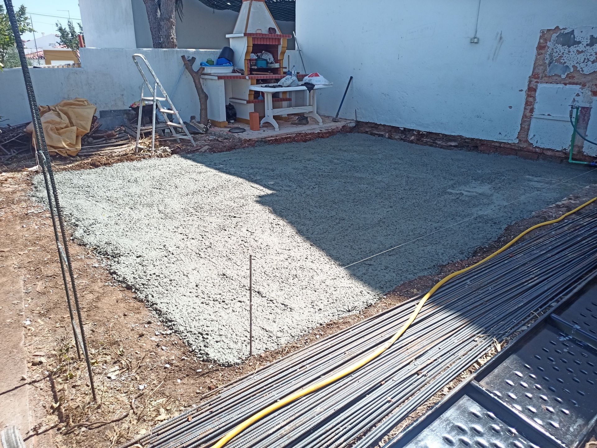 Canteiro de obras com base de concreto sendo concretada; vergalhões, ferramentas e uma parede parcialmente construída são visíveis.
