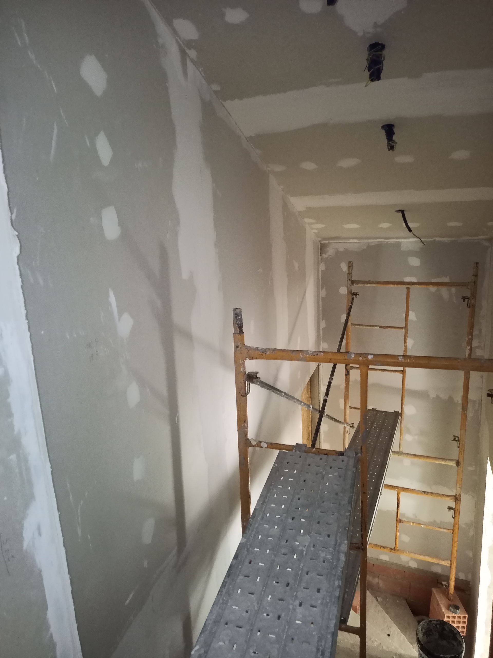 Cena interna de uma obra com paredes de gesso cartonado, andaimes e iluminação suspensa.