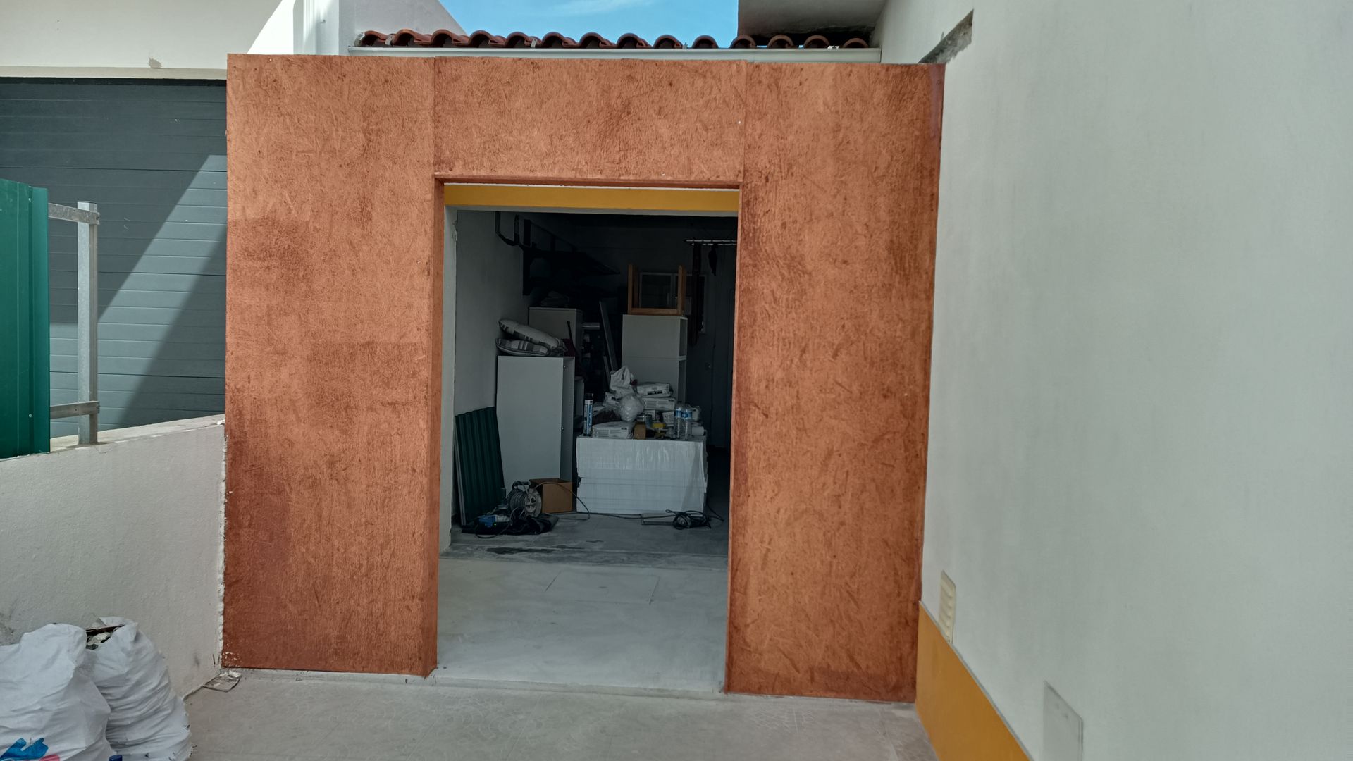 Porta aberta com moldura texturizada na cor cobre, que dá acesso a um interior desorganizado. O exterior possui paredes brancas.