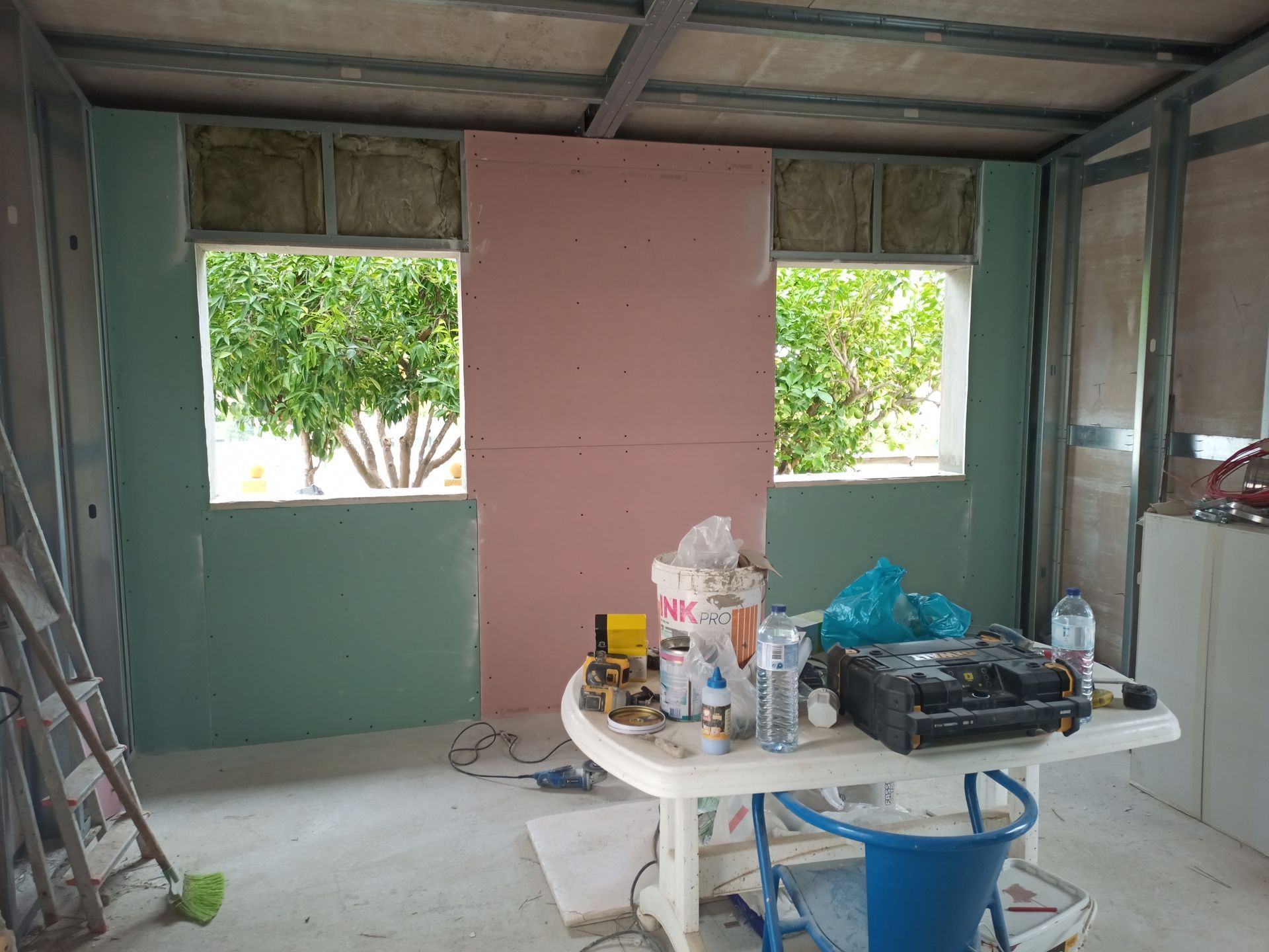 Interior de um cômodo em construção, com janelas que mostram árvores, parcialmente com paredes de gesso acartonado. Ferramentas e materiais sobre uma mesa.