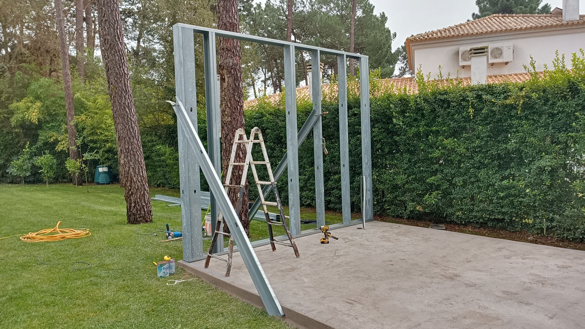 Estrutura metálica sendo construída ao ar livre sobre cascalho, com uma escada e uma cerca viva verde ao fundo.