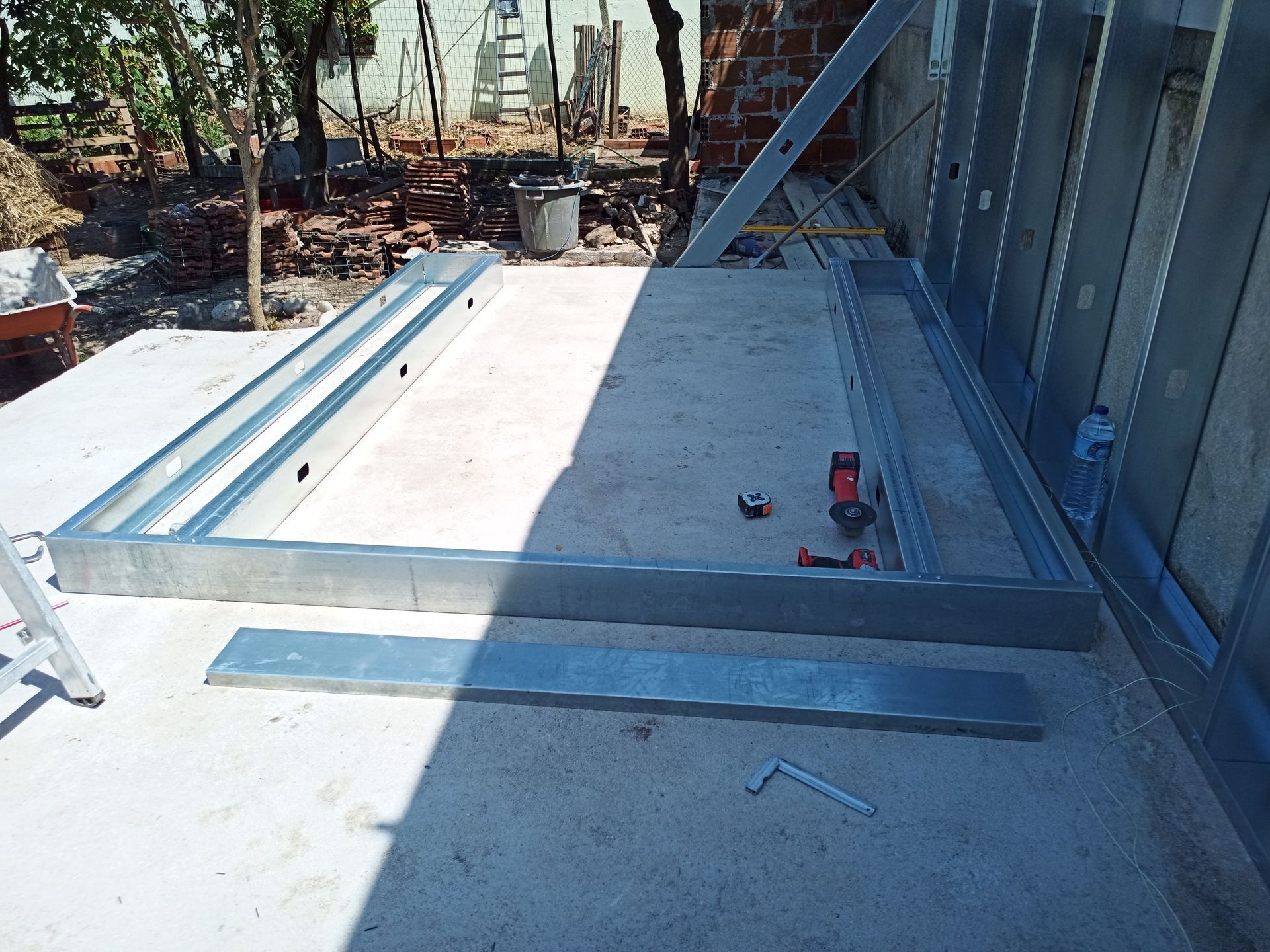 Componentes de uma estrutura metálica de porta sobre uma superfície de concreto durante a construção. Ferramentas e uma estrutura de parede também são visíveis.