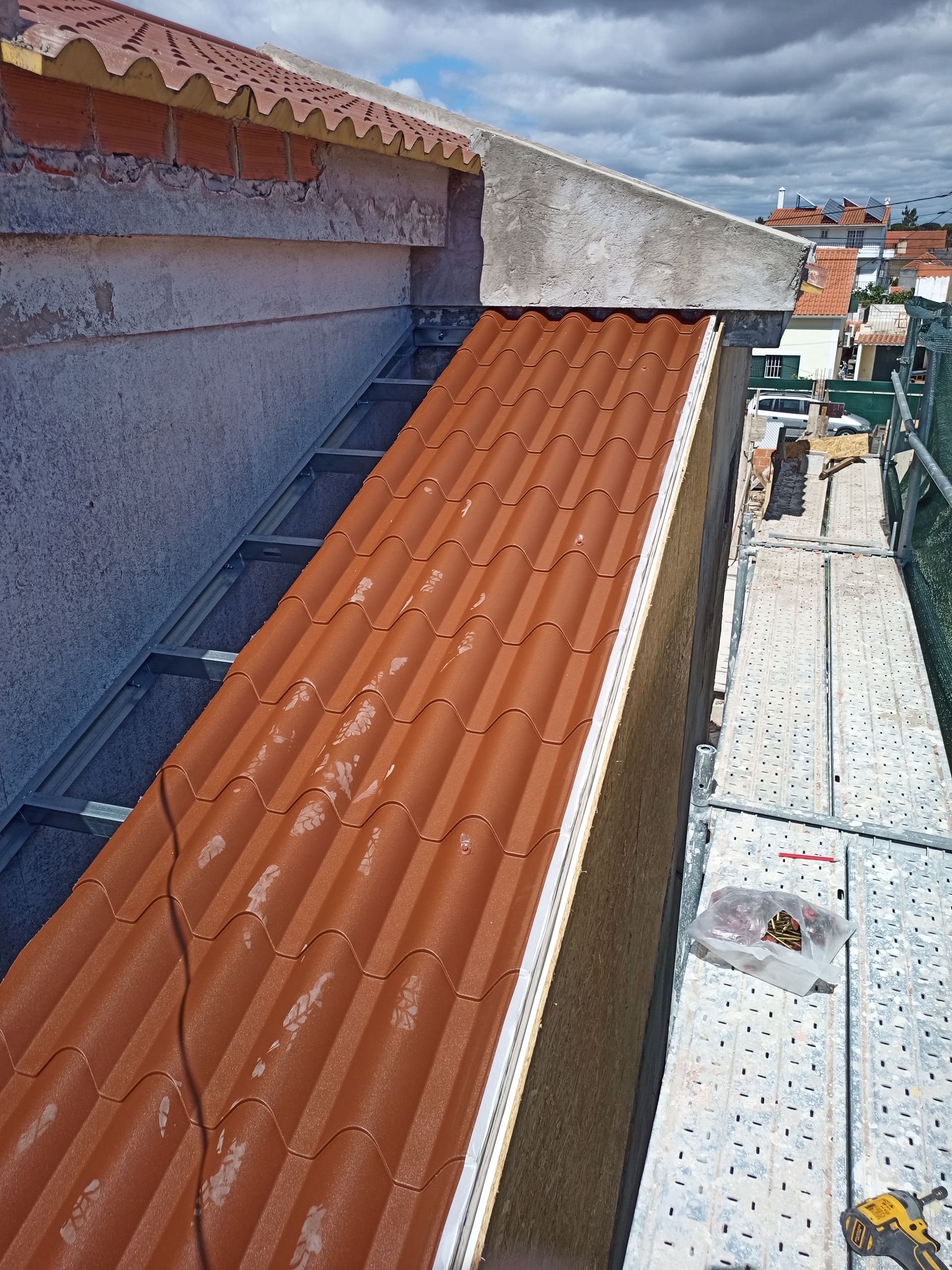 Painéis de telhado ondulados vermelhos sendo instalados perto da borda de um edifício com o auxílio de andaimes.